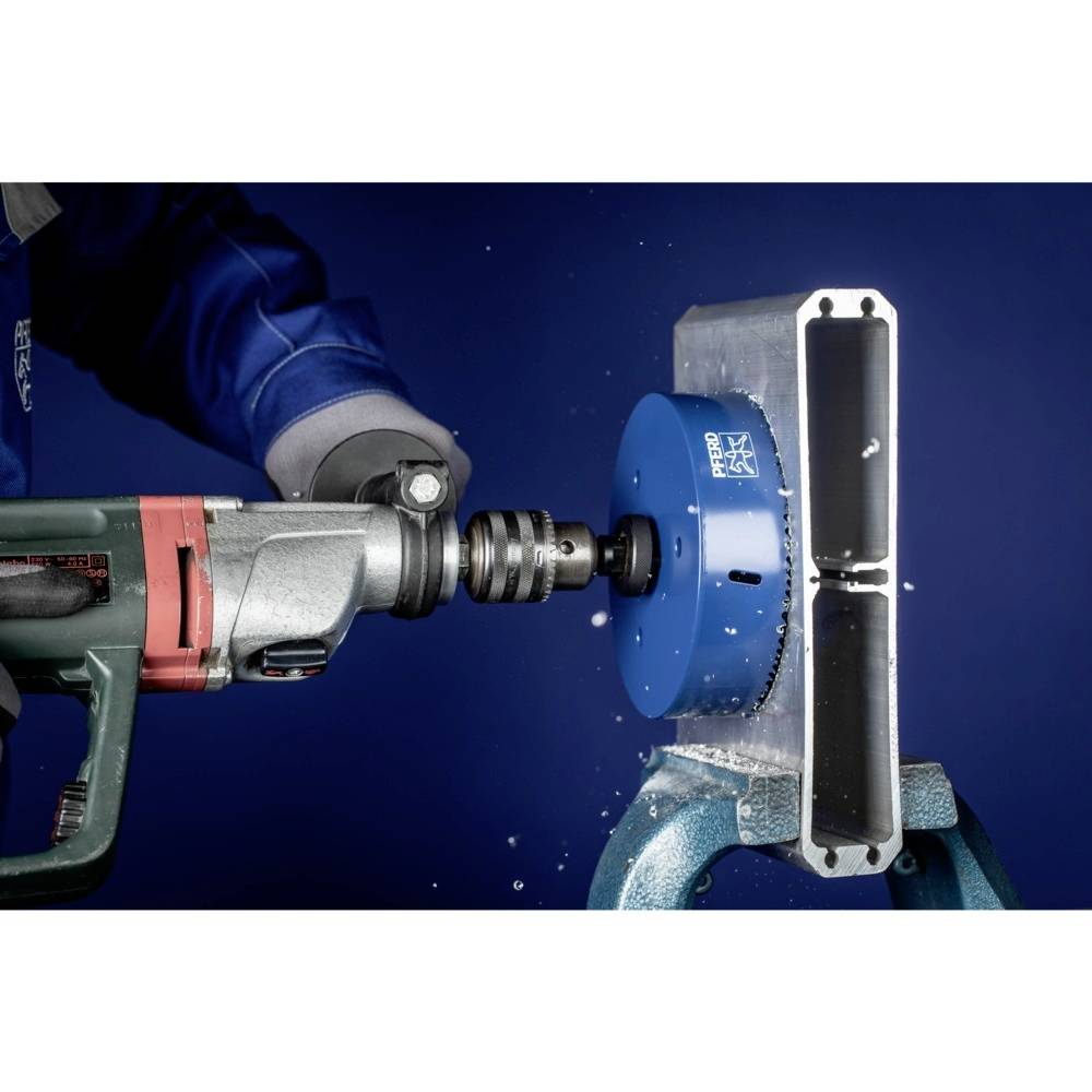 Oseba v delovni obleki z električno vrtalno napravo vrta skozi okrogel moder kovinski kos, obdan z ostružki.