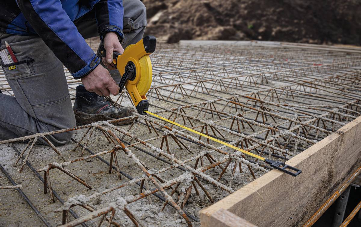 Gradbeni delavec s pomočjo merilnega traku meri armaturne palice na betonski gradbeni lokaciji. Armature so pomembne za stabilnost strukture.