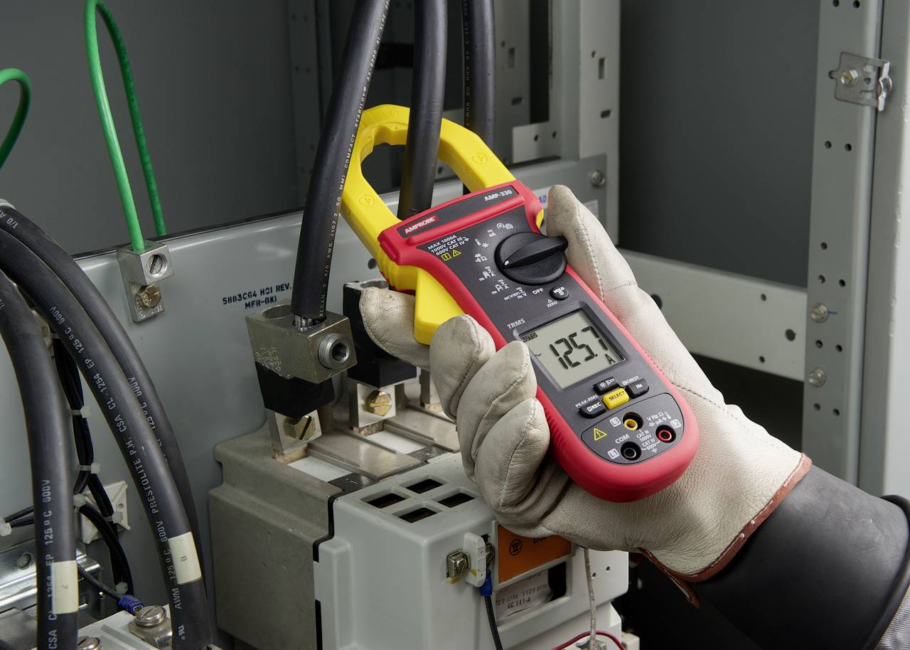 Rokavica oblečena roka drži digitalno klešče z merilnikom, ki prikazuje meritev 125. Merilnik meri električne kable v električni omarici.