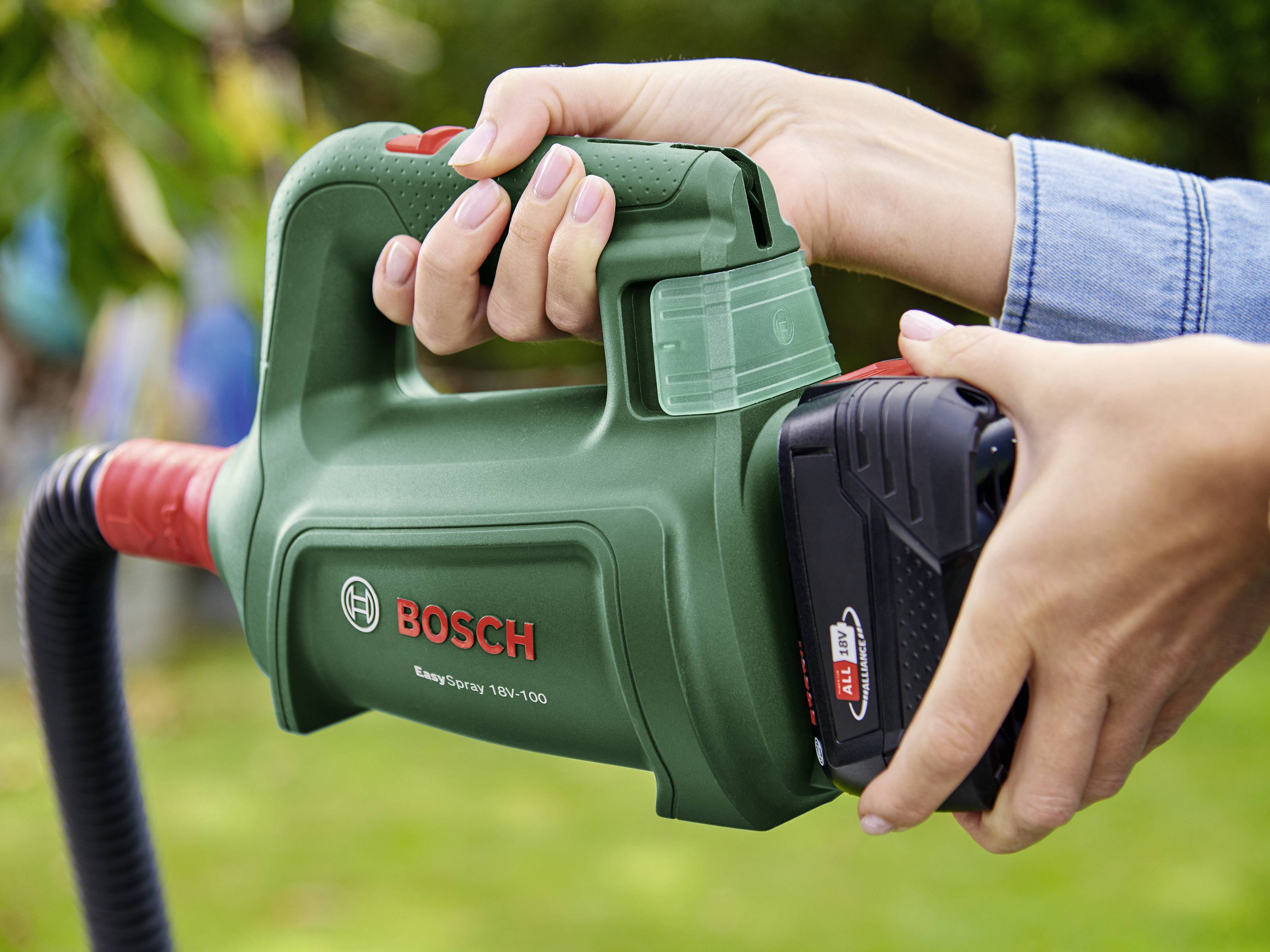 Dve roki držita zeleno vrtno orodje Bosch z akumulatorjem v vrtu. Logotip Bosch je viden; naprava je v uporabi.