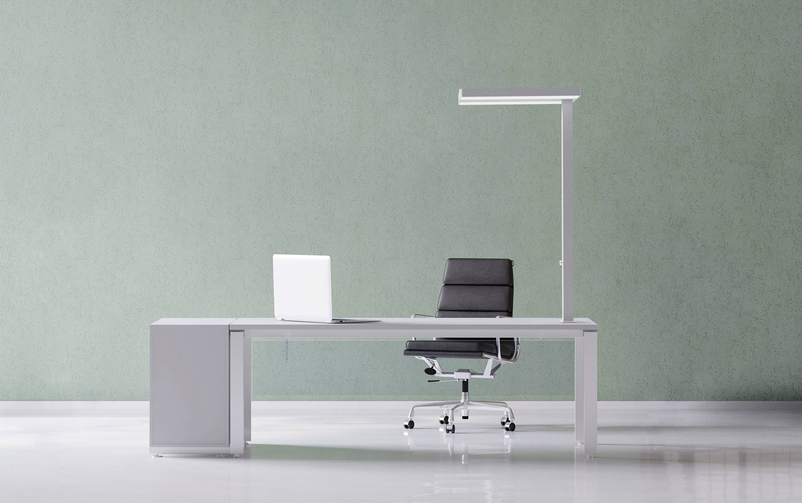 Minimalistično pisarniško delovno mesto z prenosnim računalnikom na dolgem sivem stolu, ergonomskim stolom in stoječo lučjo pred zeleno steno.