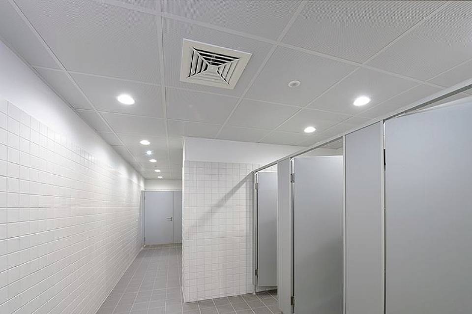 Sterilen, sodoben sanitarni prostor z belimi ploščicami, več zaprtimi vrati kabin in svetlo stropno razsvetljavo.