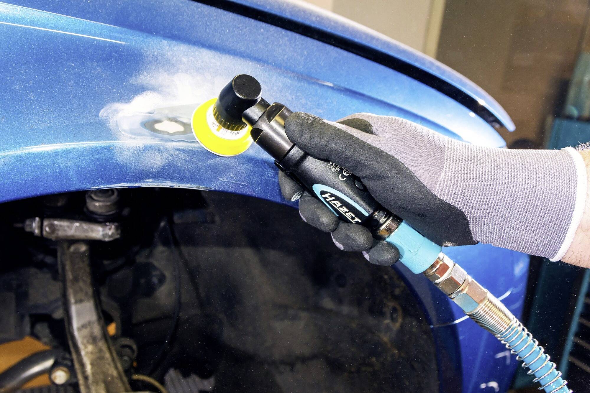 Delavec s peskanjem obdeluje modro karoserijo avtomobila. Na mestu obdelave so vidni oblaki prahu.