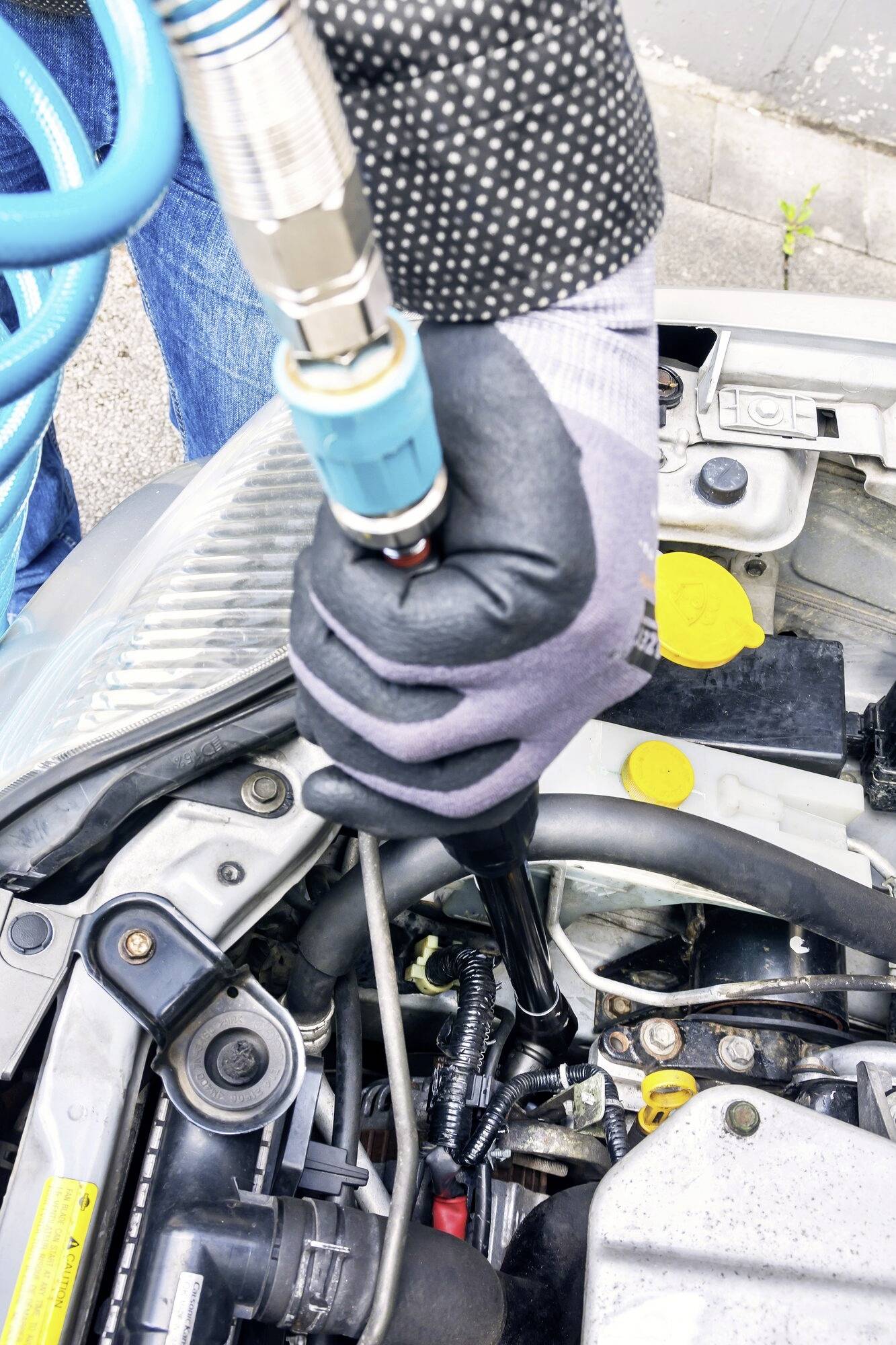 Oseba nosi rokavice in uporablja pnevmatsko orodje med delom na odprtem motorju vozila. Prizor avtomehanične delavnice.