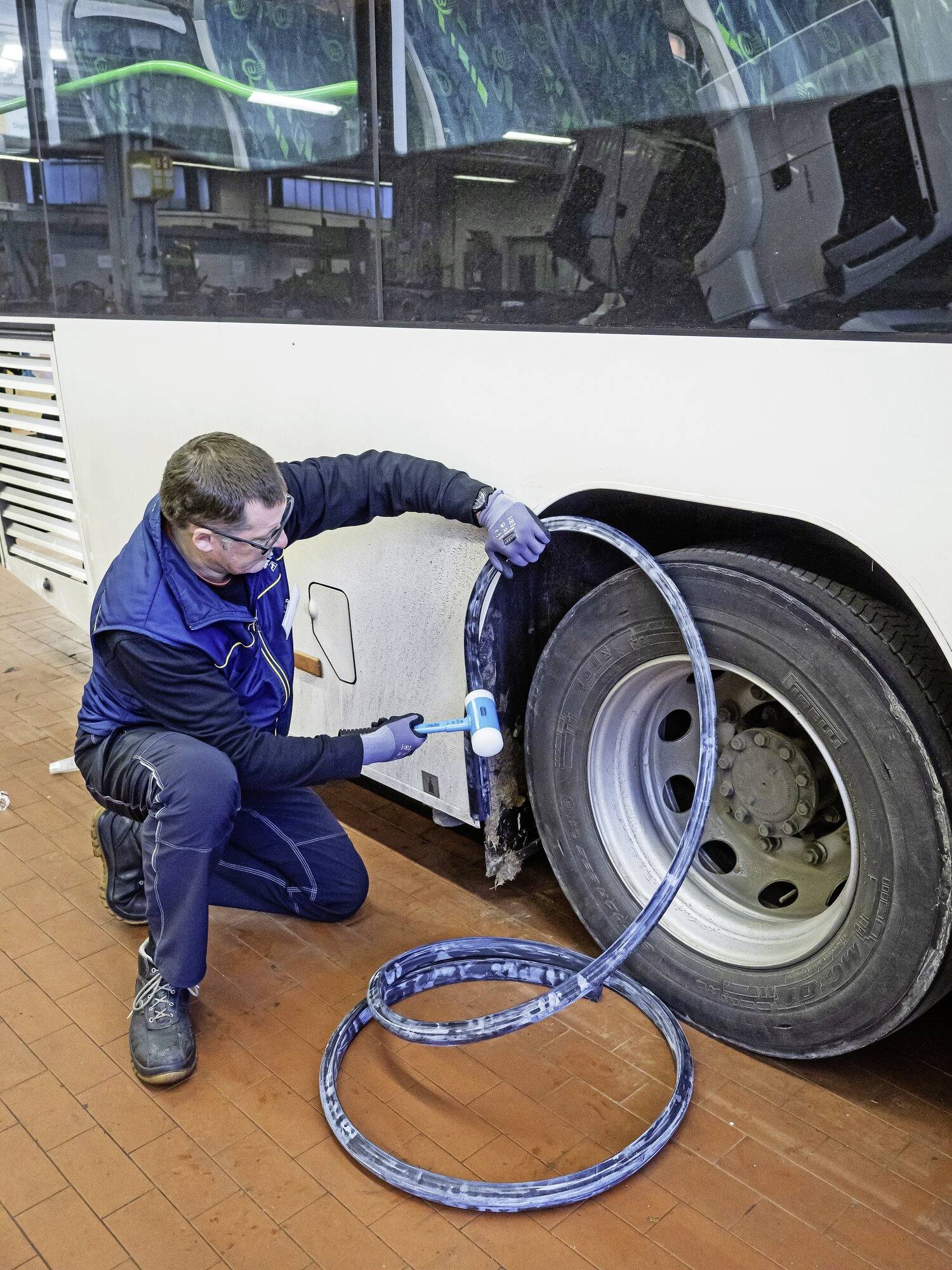 Moški klečí poleg avtobusne pnevmatike v delavnici in opravlja vzdrževalna dela. V rokah drži kladivo in cev.