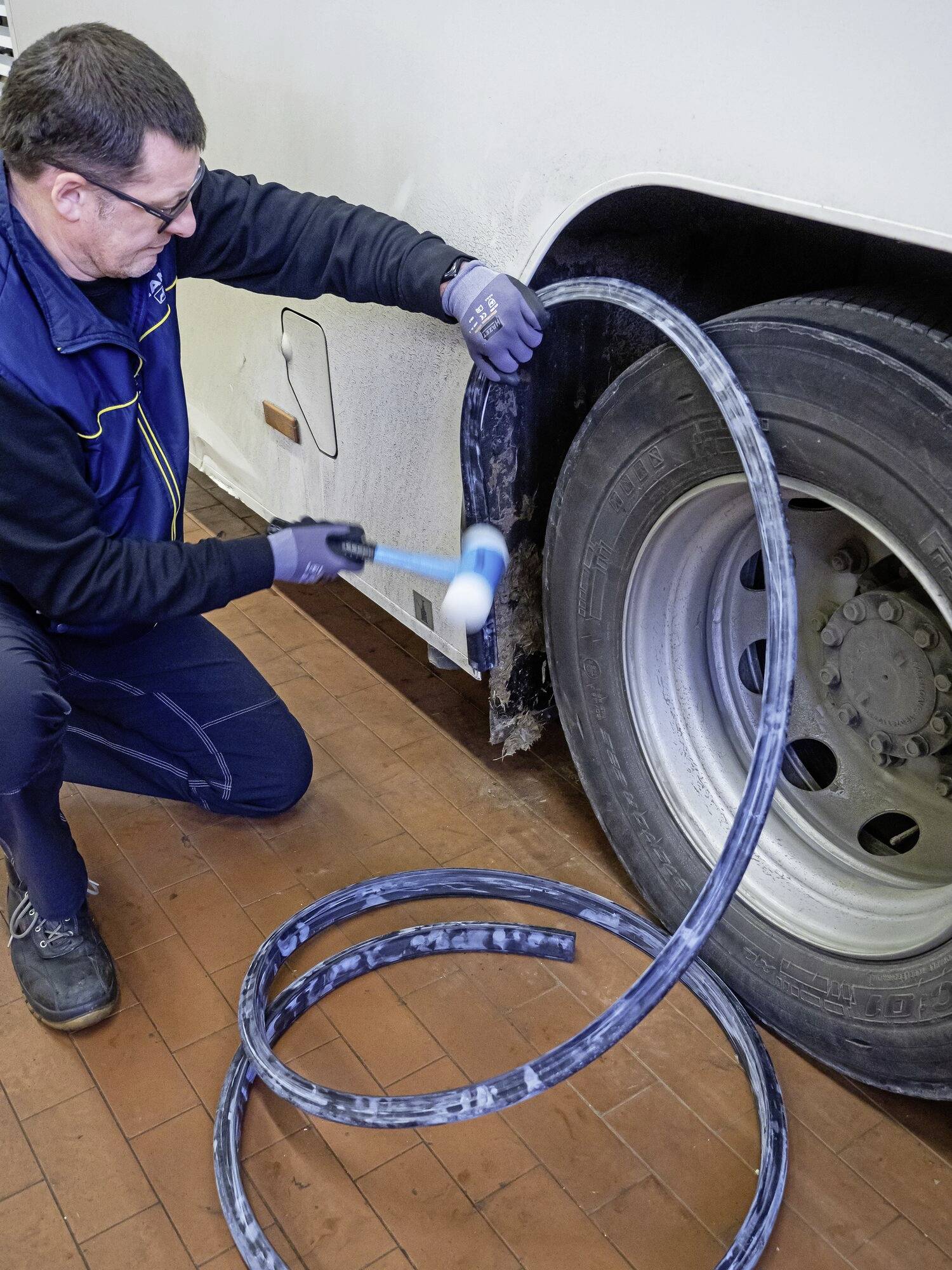 Moški popravlja avtobusnemu kolesu pnevmatiko v delavnici, medtem ko drži kladivo in orodje. Pnevmatika je že delno odstranjena.