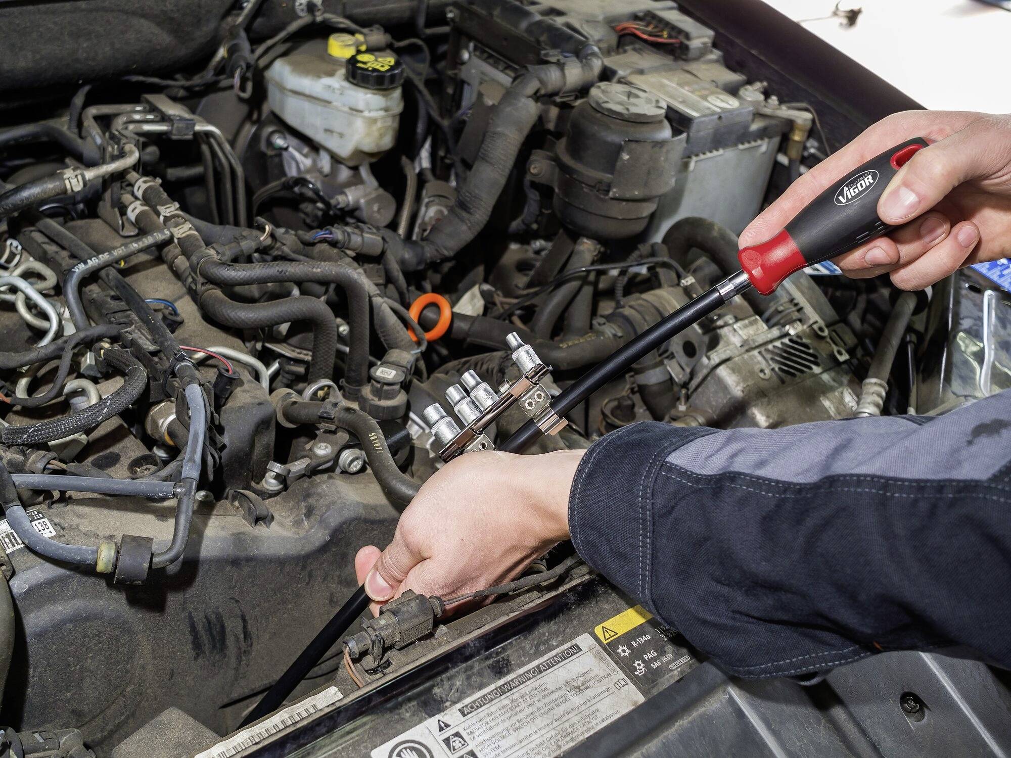 Oseba popravlja motor avtomobila s ključem. V ospredju so vidni različni kabli in deli.