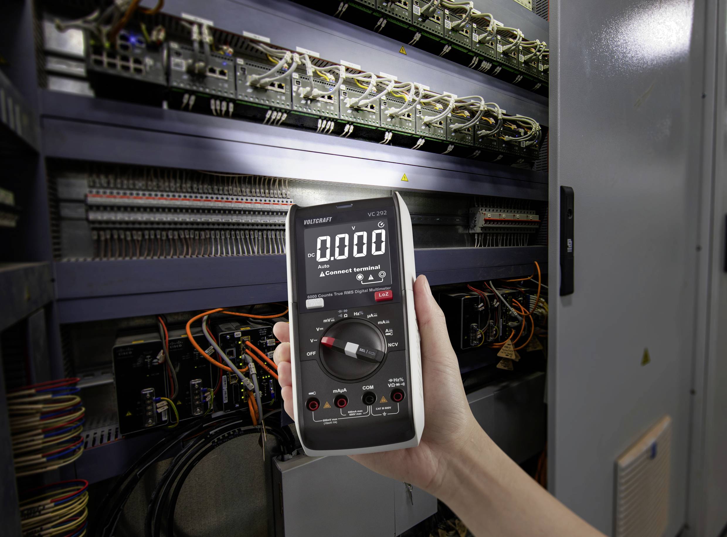 Roka drži digitalni multimeter, nastavljen na 0,000, pred odprto električno omarico, polno kablov in elektronskih komponent.