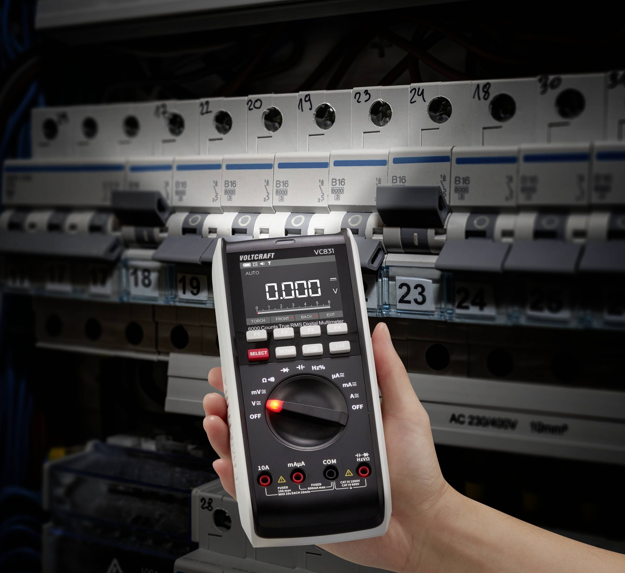 Roka drži digitalni multimeter pred odprtim omarico z varovalkami in več instalacijskimi odklopniki.