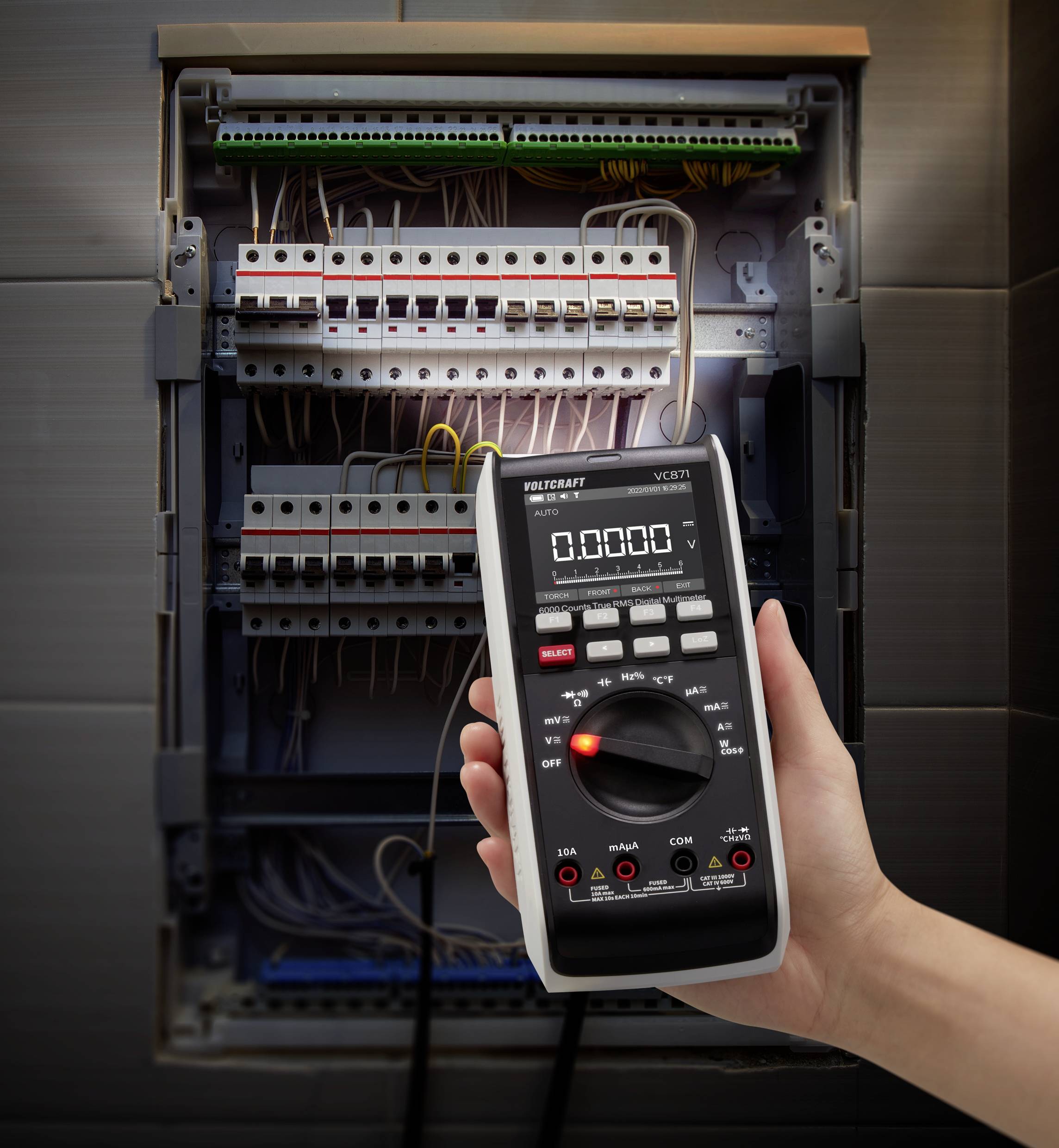 Roka drži digitalni multimeter pred odprtim elektroenergetskim razdelilnikom, da bi izmerila električne vrednosti.