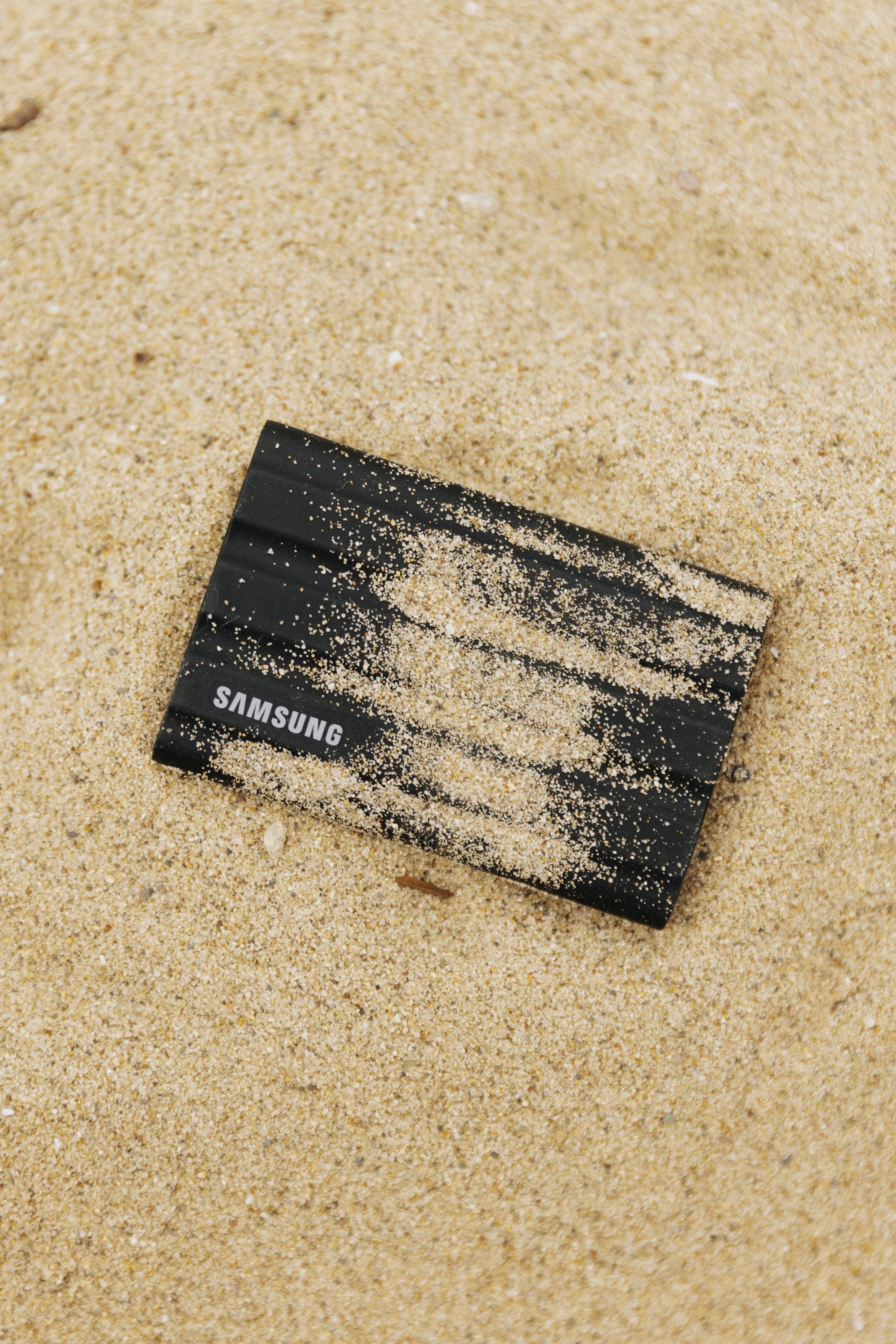 Črn Samsung zunanji trdi disk leži v pesku. Zgoraj je delno prekrit s peskom.
