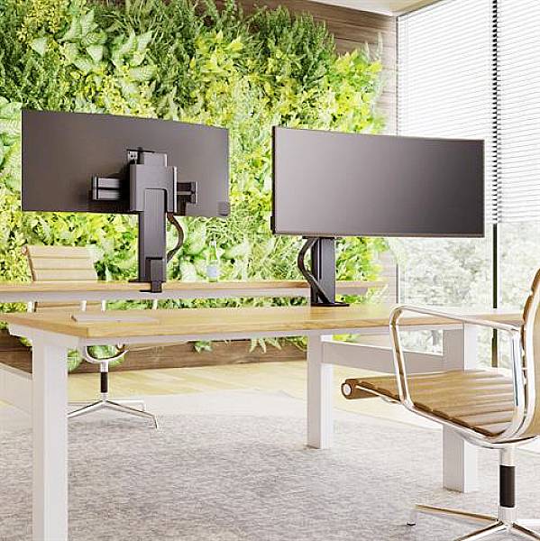 Sodoben delovni prostor z dvema monitorjema, leseno mizo in ergonomskim stolom. V ozadju zelena stena z rastlinami.