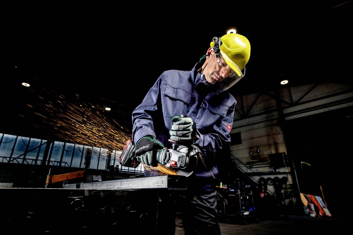 Delavec v zaščitni opremi in čeladi z kotnimbrusilnikom brusi kovino, iskre letijo v industrijskem okolju.