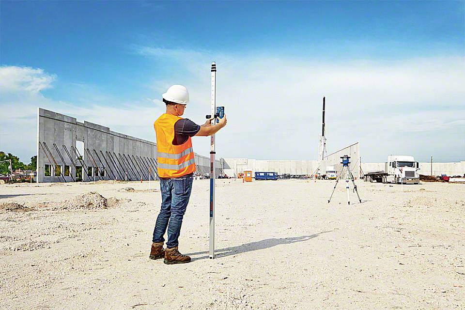 Gradbeni delavec v zaščitni opremi meri prazno gradbišče z laserskim merilnikom. V ozadju so betonske stene in oprema.