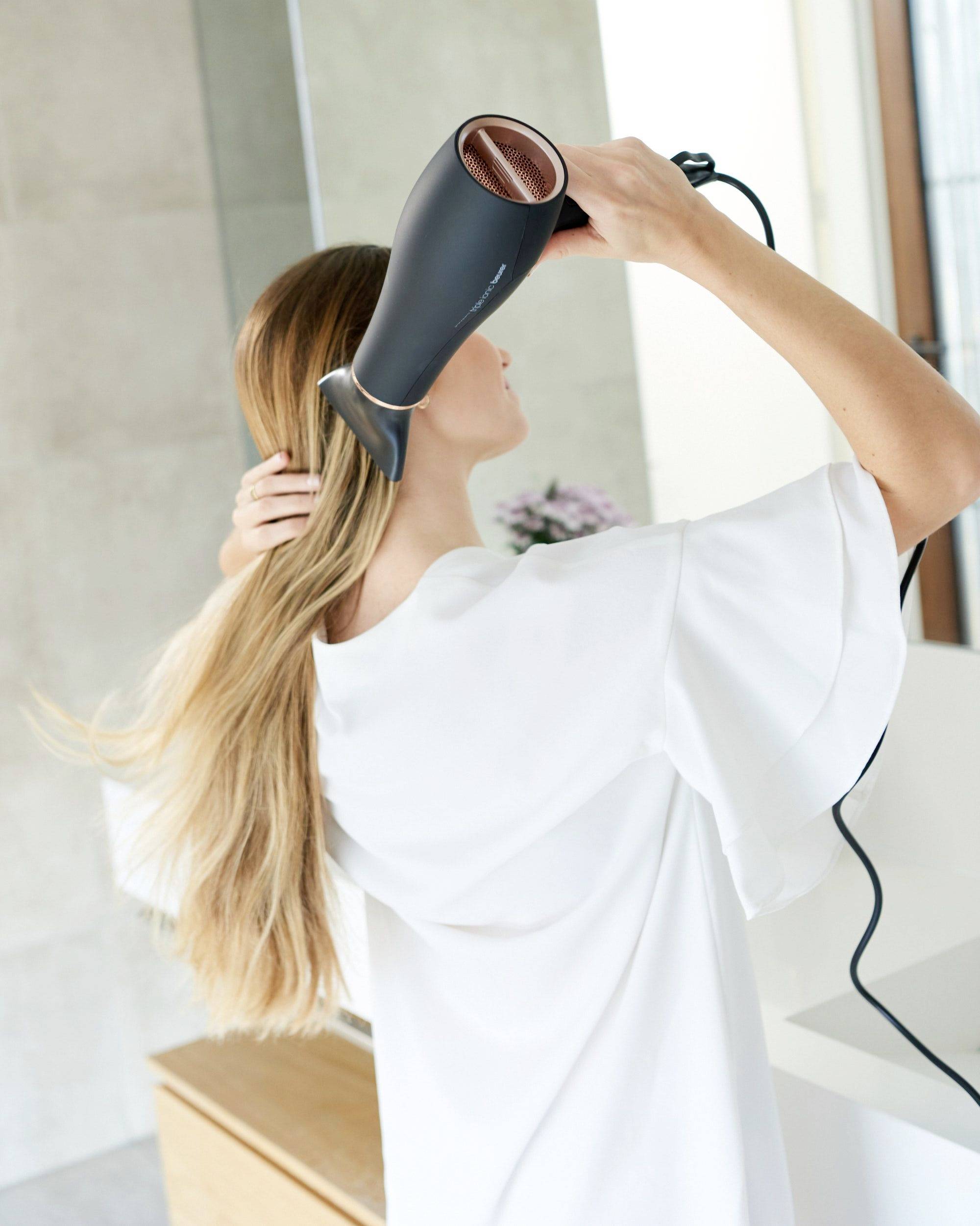 Ženska z dolgimi, svetlimi lasmi s sušilnikom suši lase v kopalnici. Oblečena je v belo zgornjo oblačilo. V ozadju so nejasno vidni cvetovi.