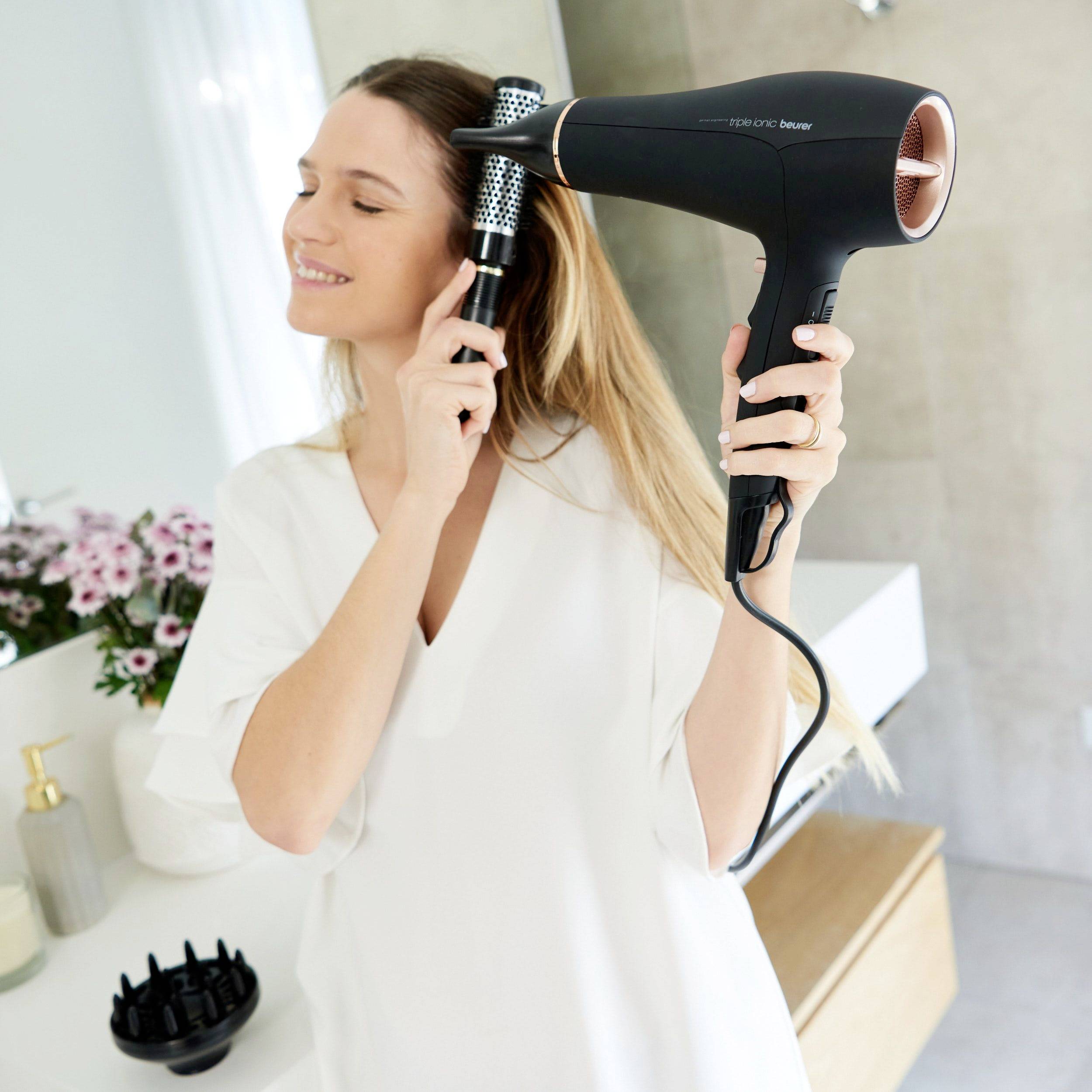 Oseba si suši lase s črnim sušilnikom las v svetli kopalnici. V ozadju so vidni cvetje in kopalniški pripomočki.