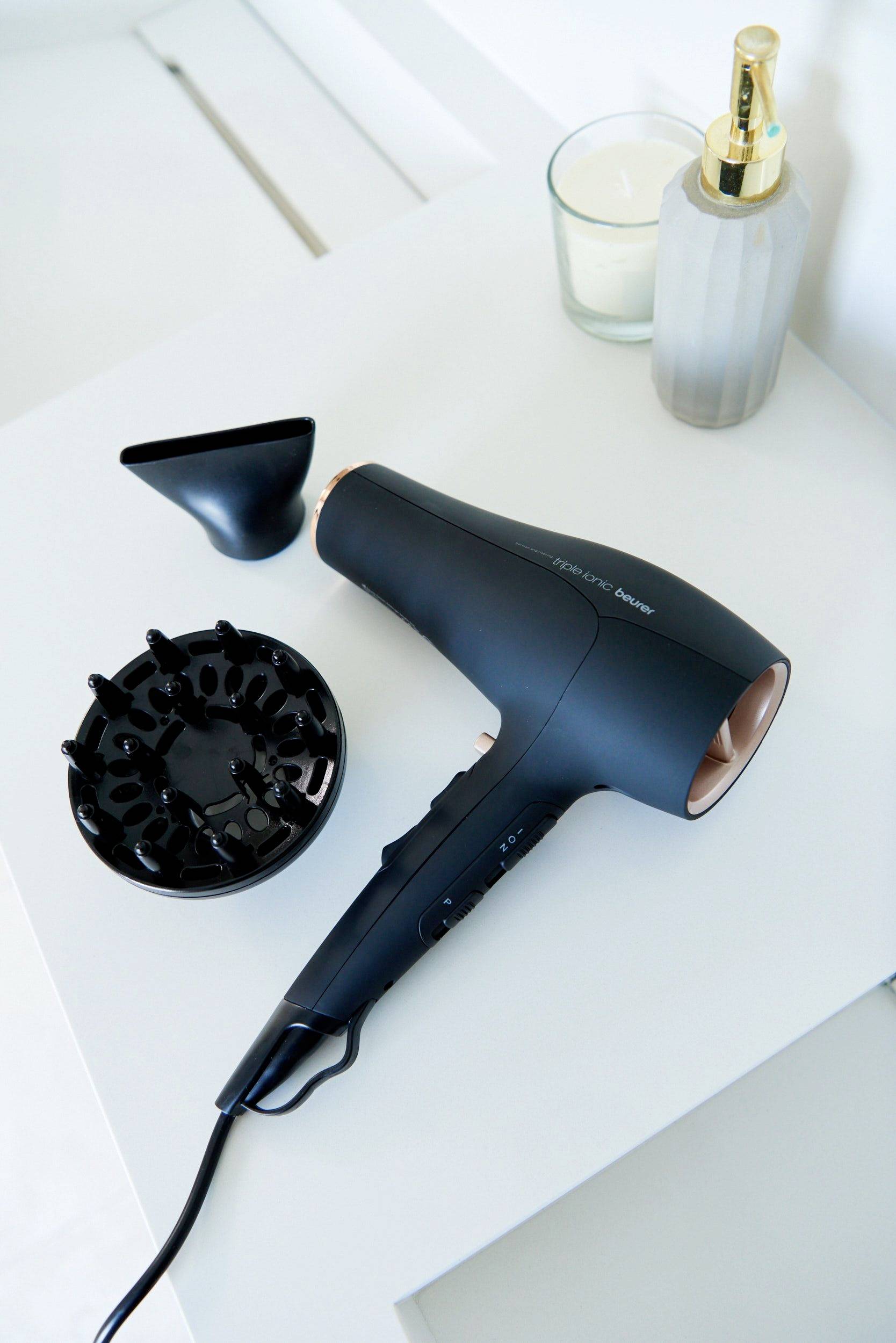 Črn sušilnik las z difuzorjem in šobo na beli mizi poleg sveče in dekorativne vaze.