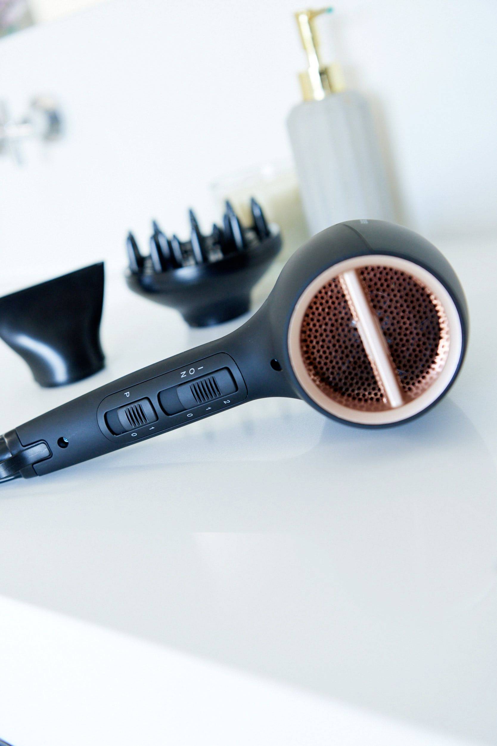 Črn sušilnik las z bakrovimi detajli leži zraven črnega difuzorja in steklenice za tekoče milo na beli mizi.