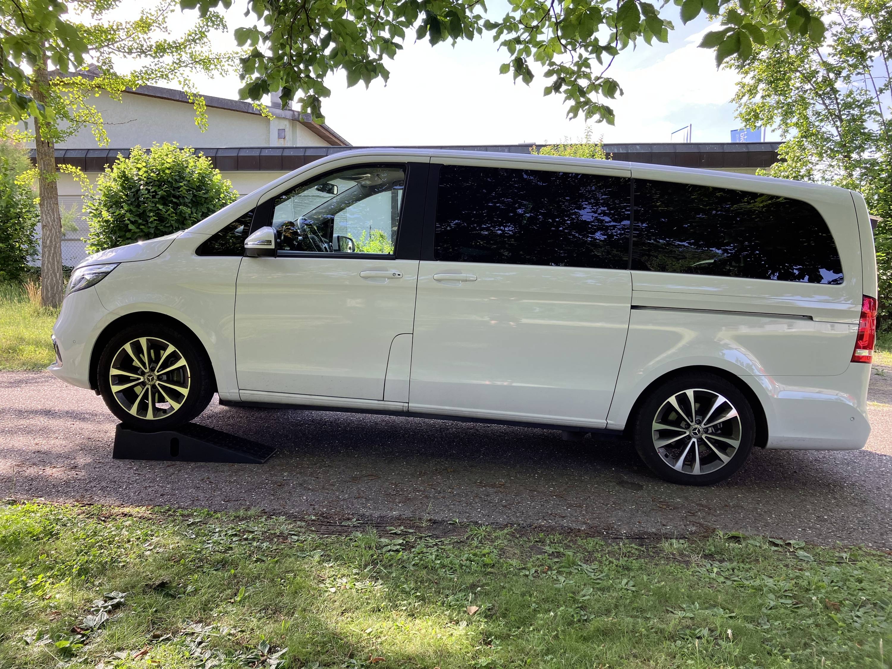 Bel kombi stoji na dovozni poti, obkrožen z drevesi in travnato površino. V ozadju so vidne nekatere stavbe.