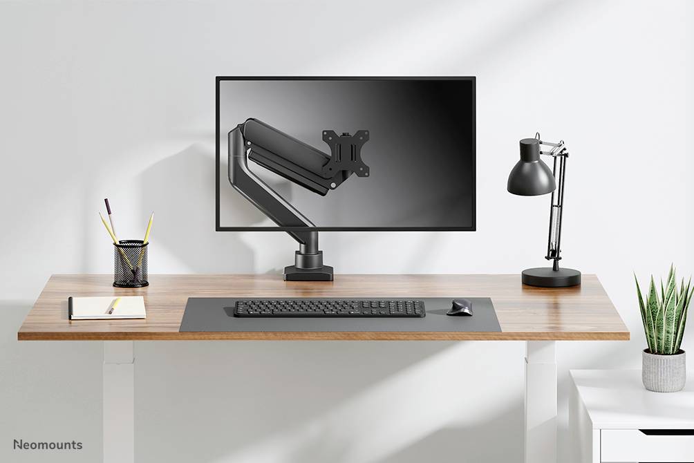 Nastavljiv pisalni mizi z regulacijo višine, nosilcem za monitor, monitorjem, tipkovnico, lučko in držalom za pisala; minimalistična delovna soba.