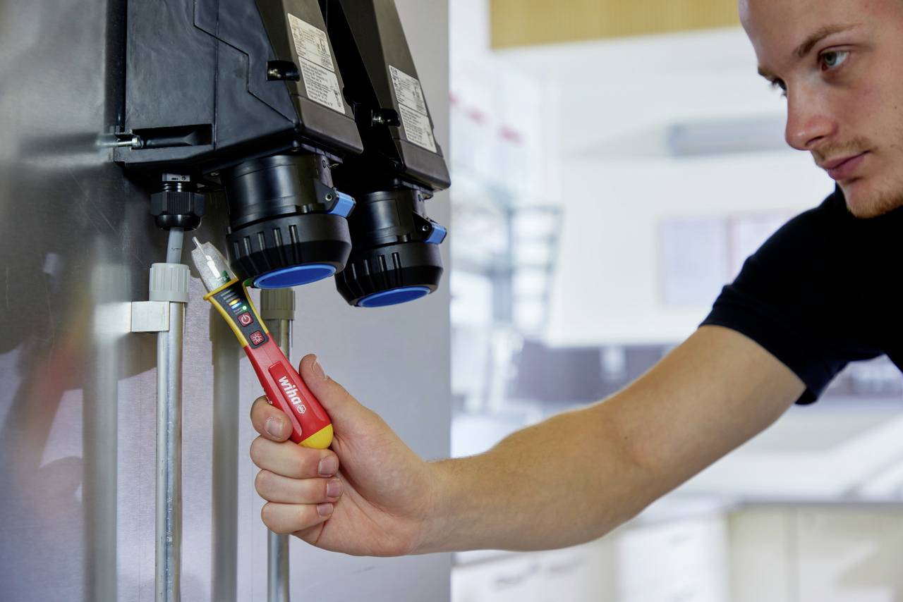 Moški s preizkuševalnikom napetosti pregleduje električno napravo. Njegov izraz je zbran.