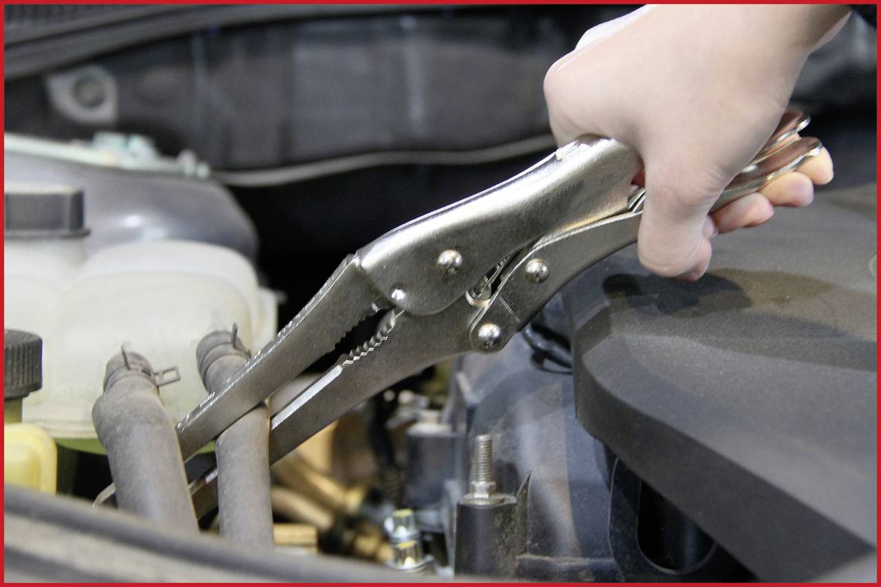 Oseba uporablja kovinske klešče za držanje ali popravilo cevi v motornem prostoru vozila.