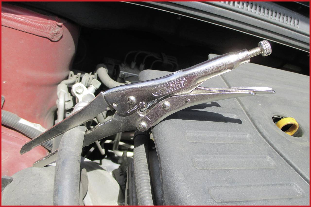 Pritrdilna klešča stiska cev v motornem prostoru avtomobila, verjetno za popravilo ali vzdrževalna dela.