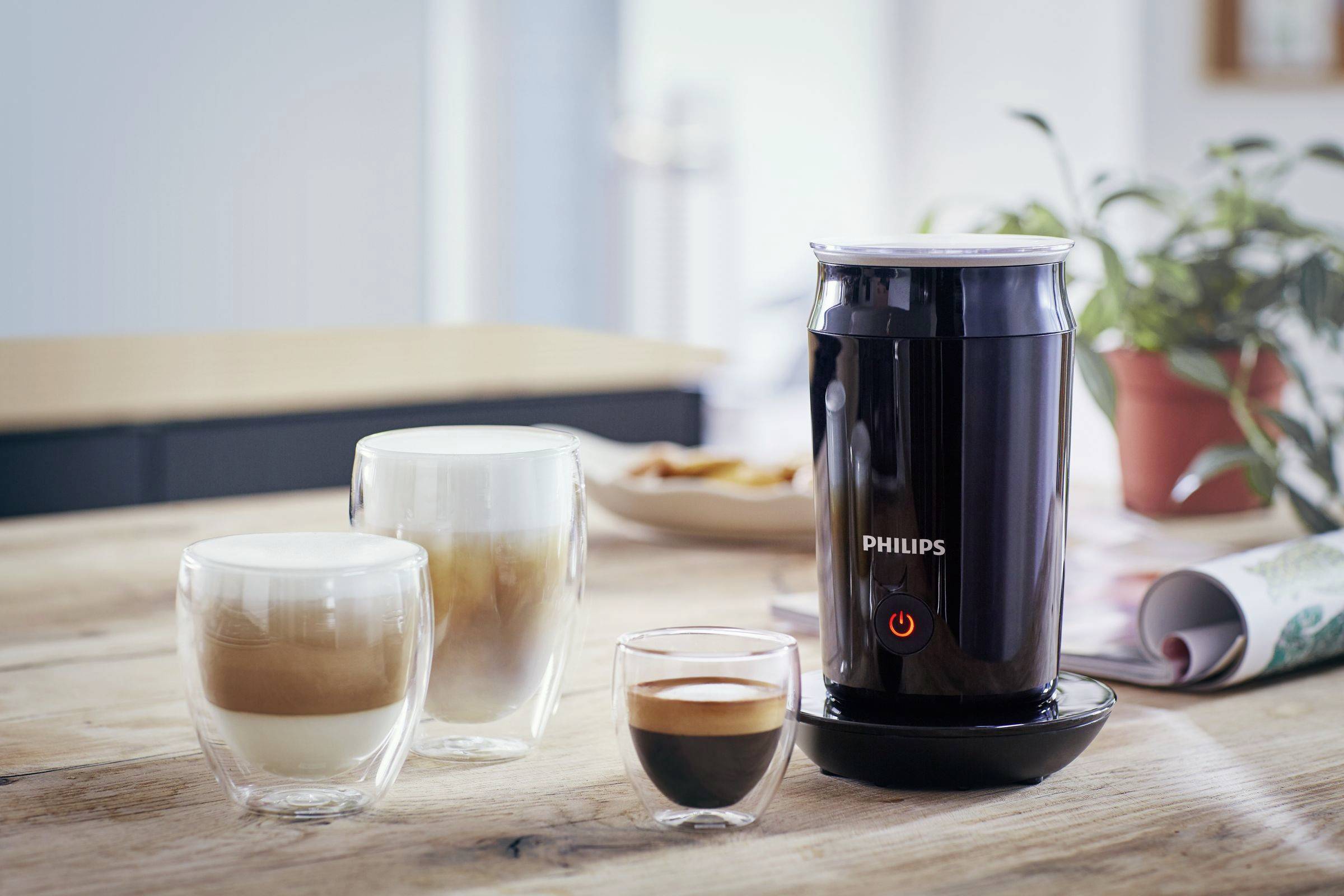 'Philipsov penilec mleka na leseni mizi, obdan s cappuccinom in espressom. V ozadju zamegljen prostor z rastlino in časopisom.'