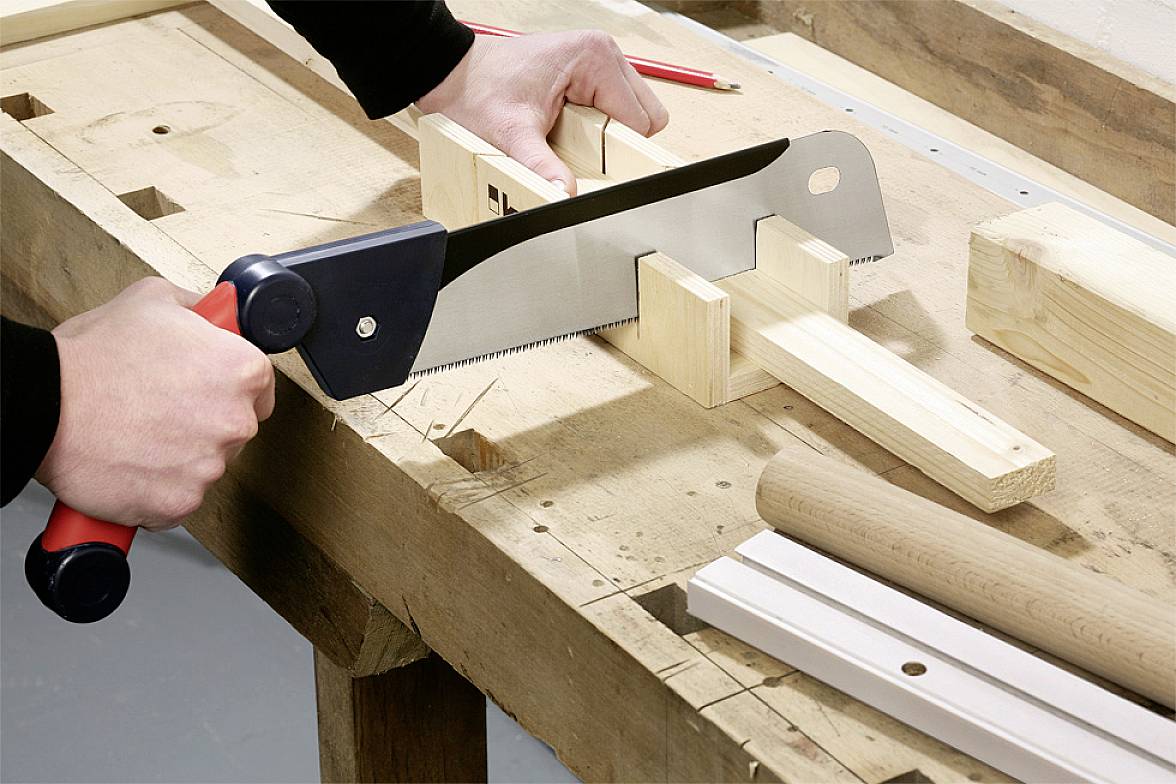 Oseba žaga les z ročno žago na delovni mizi. Okoli so leseni koščki in orodja, ki se uporabljajo za obdelavo lesa.