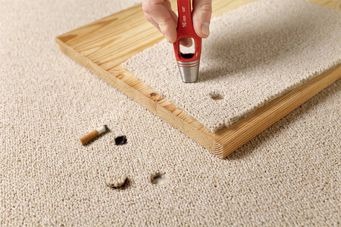 Roka uporablja tepih rezalnik na kosu preproge na leseni podlagi, da bi izrezala okrogel kos. V ozadju je cigareta in koščki zažganih lukenj.