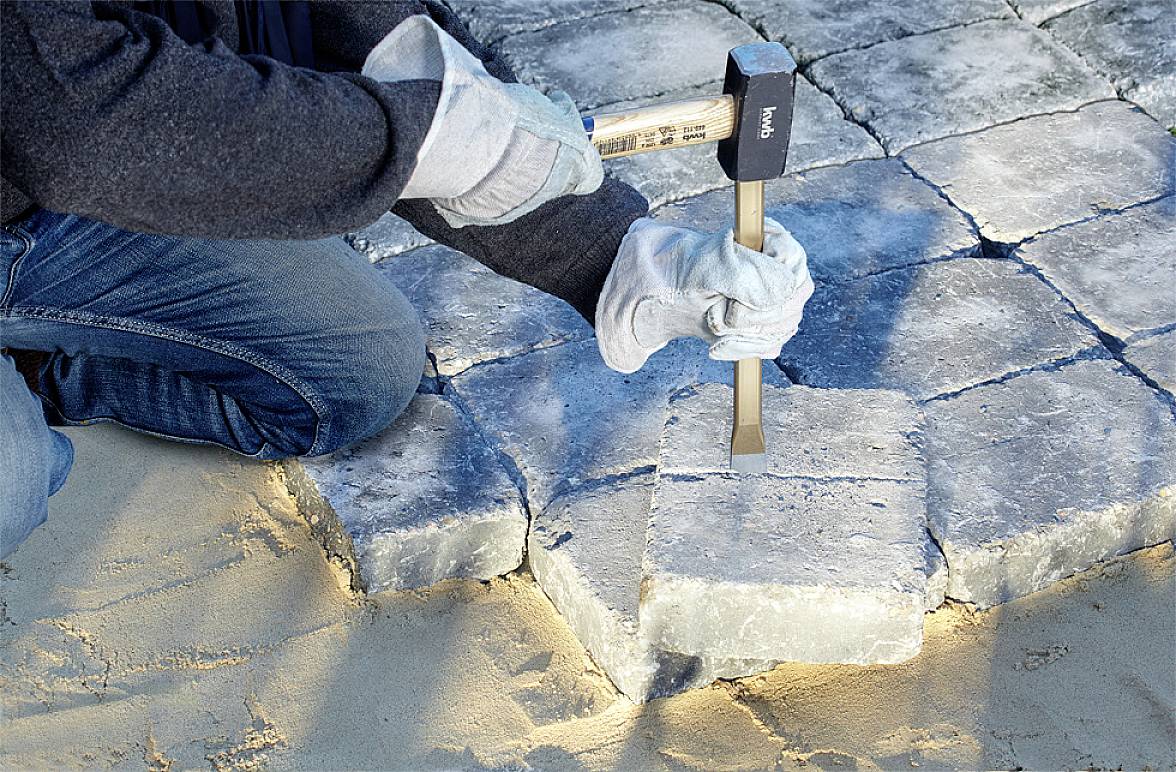 Oseba z kladivom in dleti oblikuje tlakovce. Kamni ležijo na peščeni podlagi.