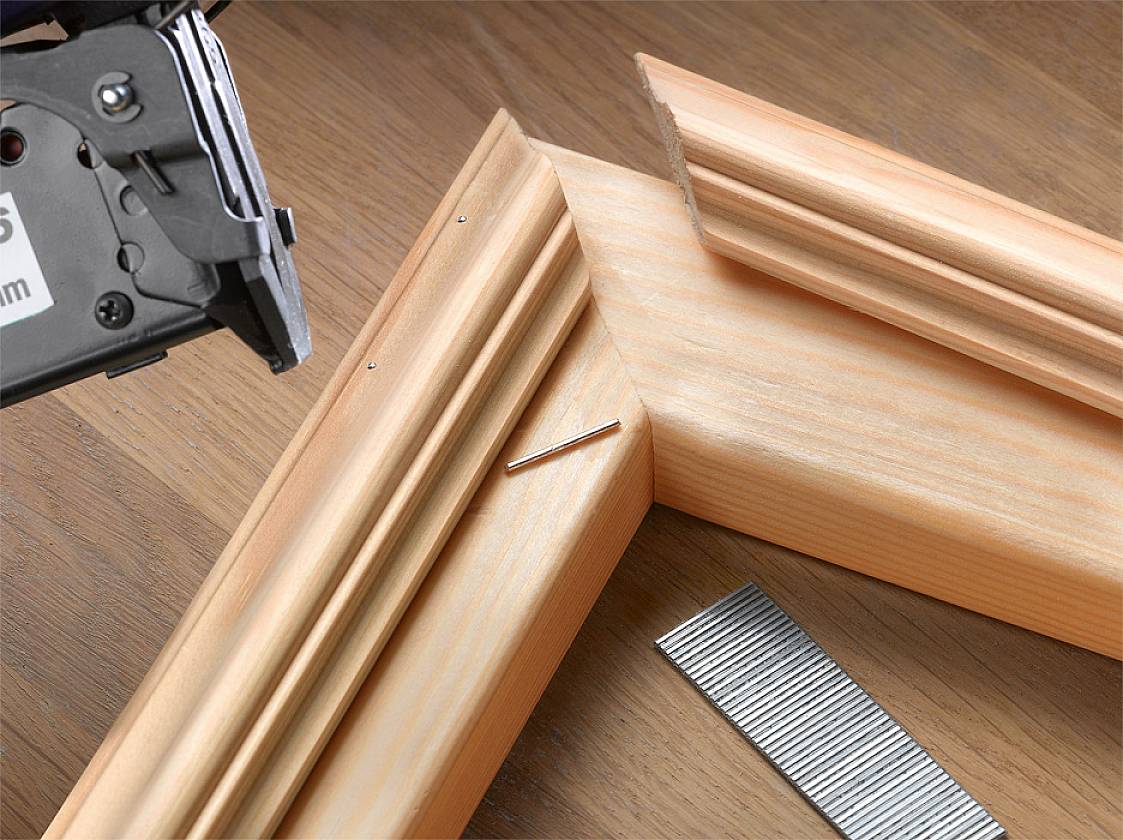 Kotna žaga reže dva lesena profila za izdelavo okvirja. Žebelj in trak žebljev ležita zraven.