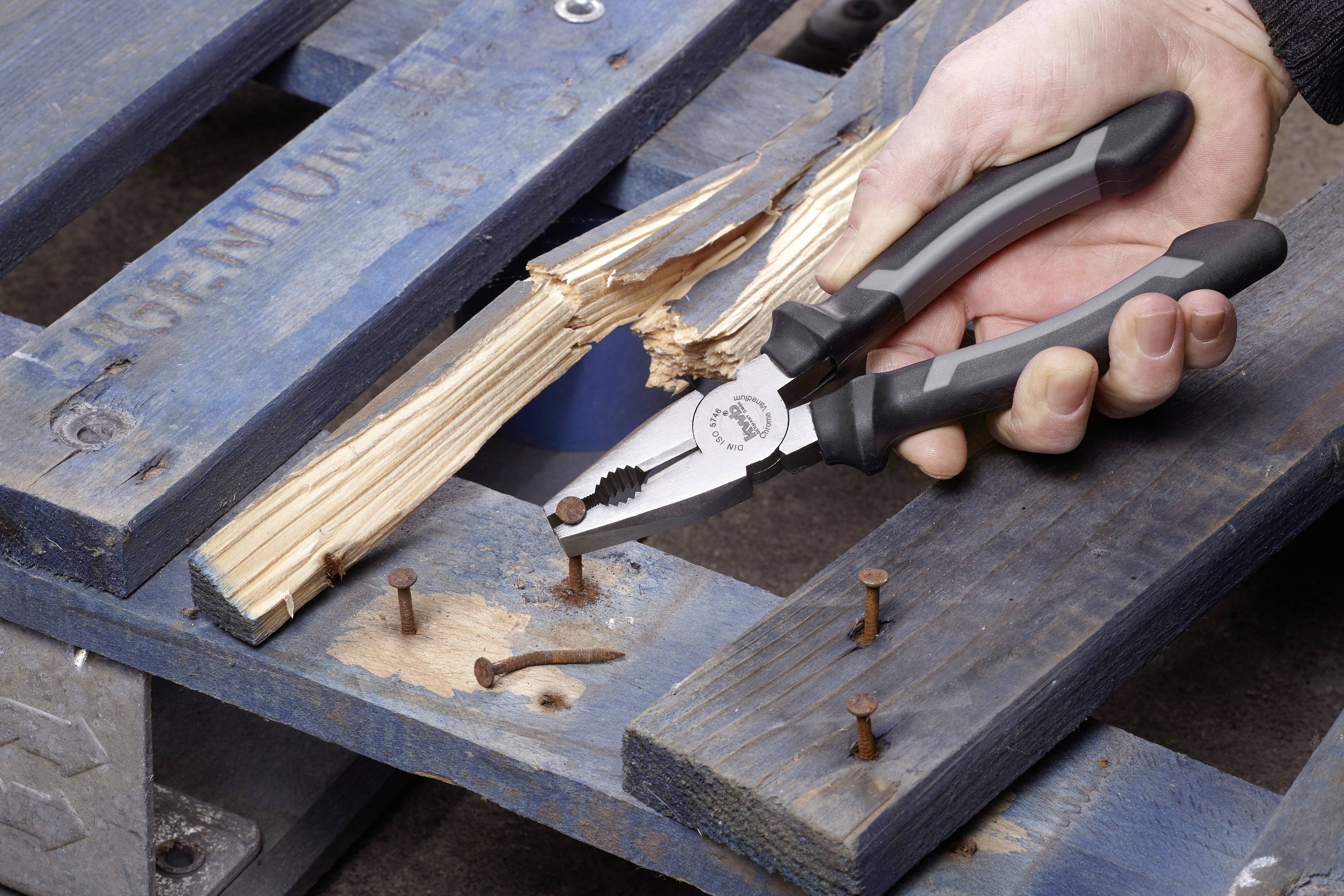 Ena roka drži klešče, da bi odstranila upognjen žebelj iz poškodovane lesene palete. Paleta je pobarvana modro.