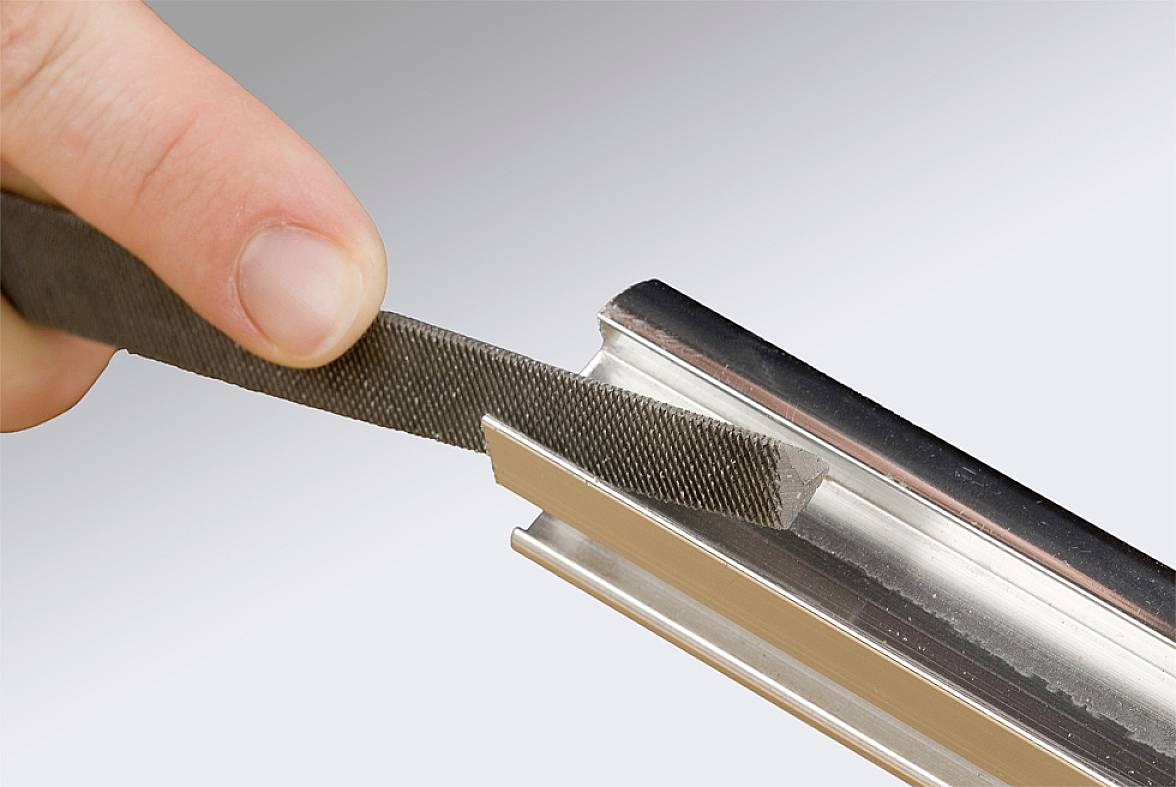 Roka s pilo obdeluje kovinski rob. Fotografija prikazuje uporabo pile za glajenje ali oblikovanje kovinskih površin.