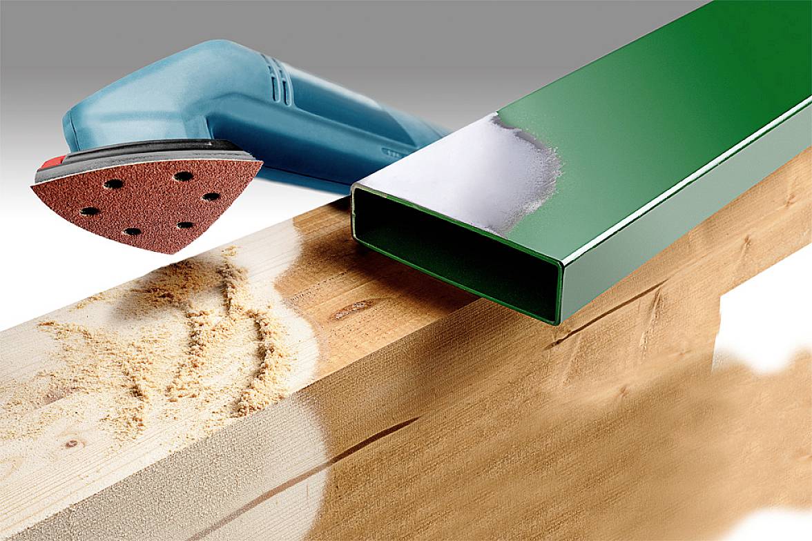 Električni brusilnik z rdečo brusilno glavo poleg obdelanega lesnega bloka in gladke zelene kovinske cevi, prikazuje učinek brušenja.