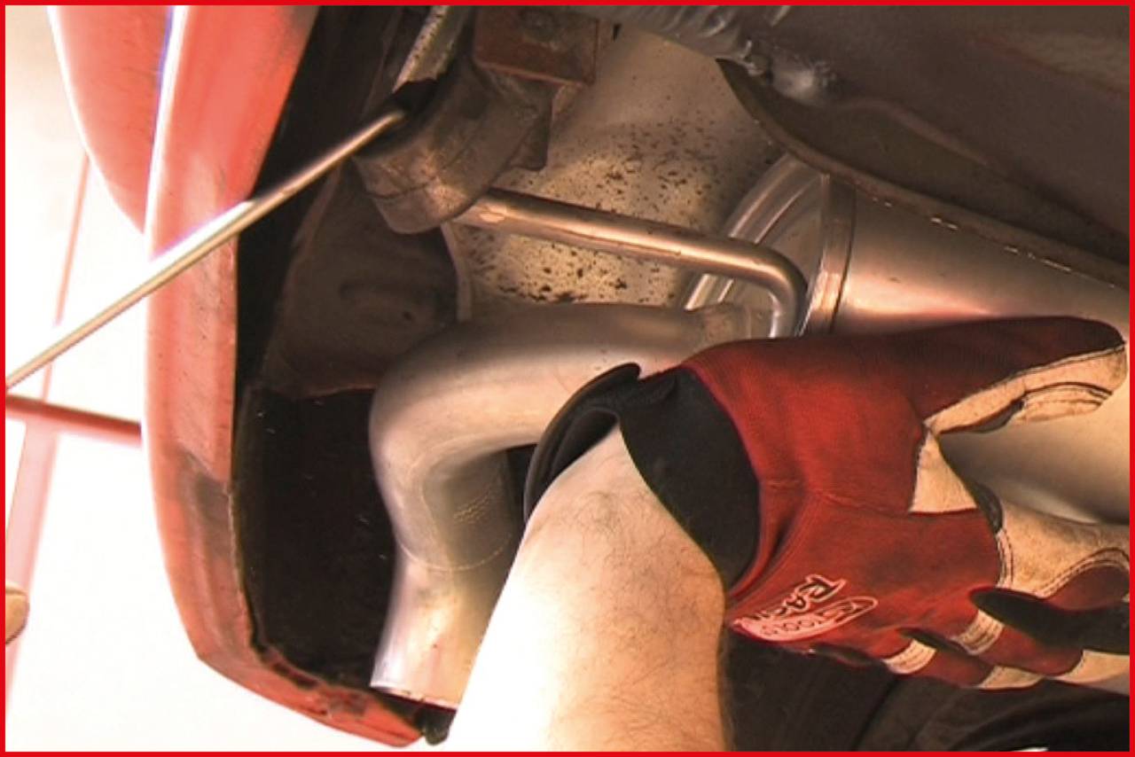 Mehanik z rdečimi rokavicami dela na izpušni cevi pod avtomobilom. V ozadju so vidni deli izpuha.