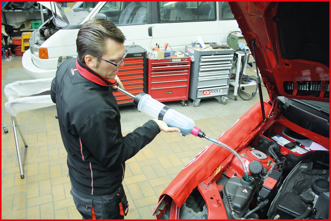 Mehanik naliva hladilno tekočino v motor rdečega avtomobila. V ozadju stojijo orodne omare in bel kombi.