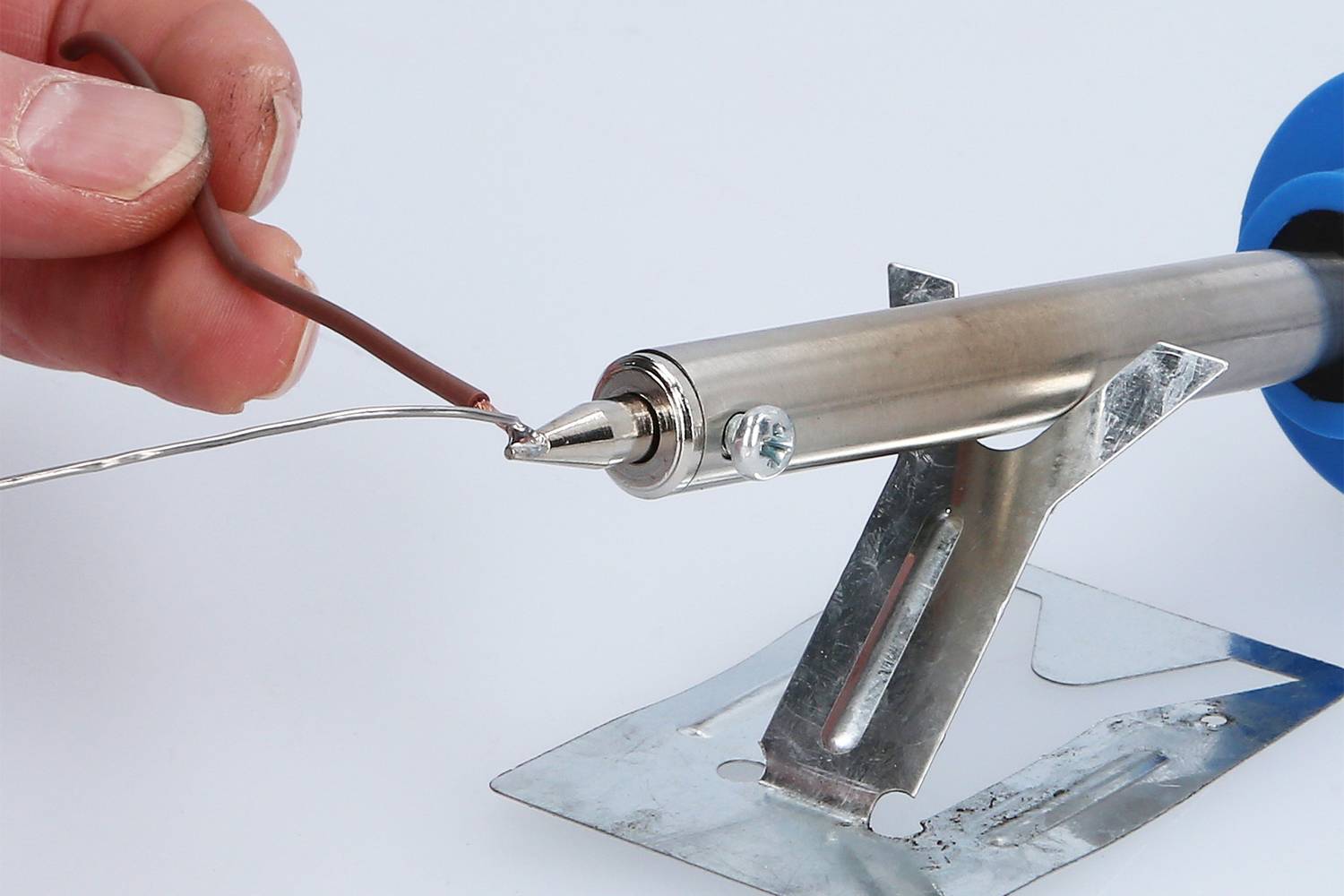 Ena roka drži rjav kabel na vroči spajkalni konici, ki počiva na kovinskem stojalu.