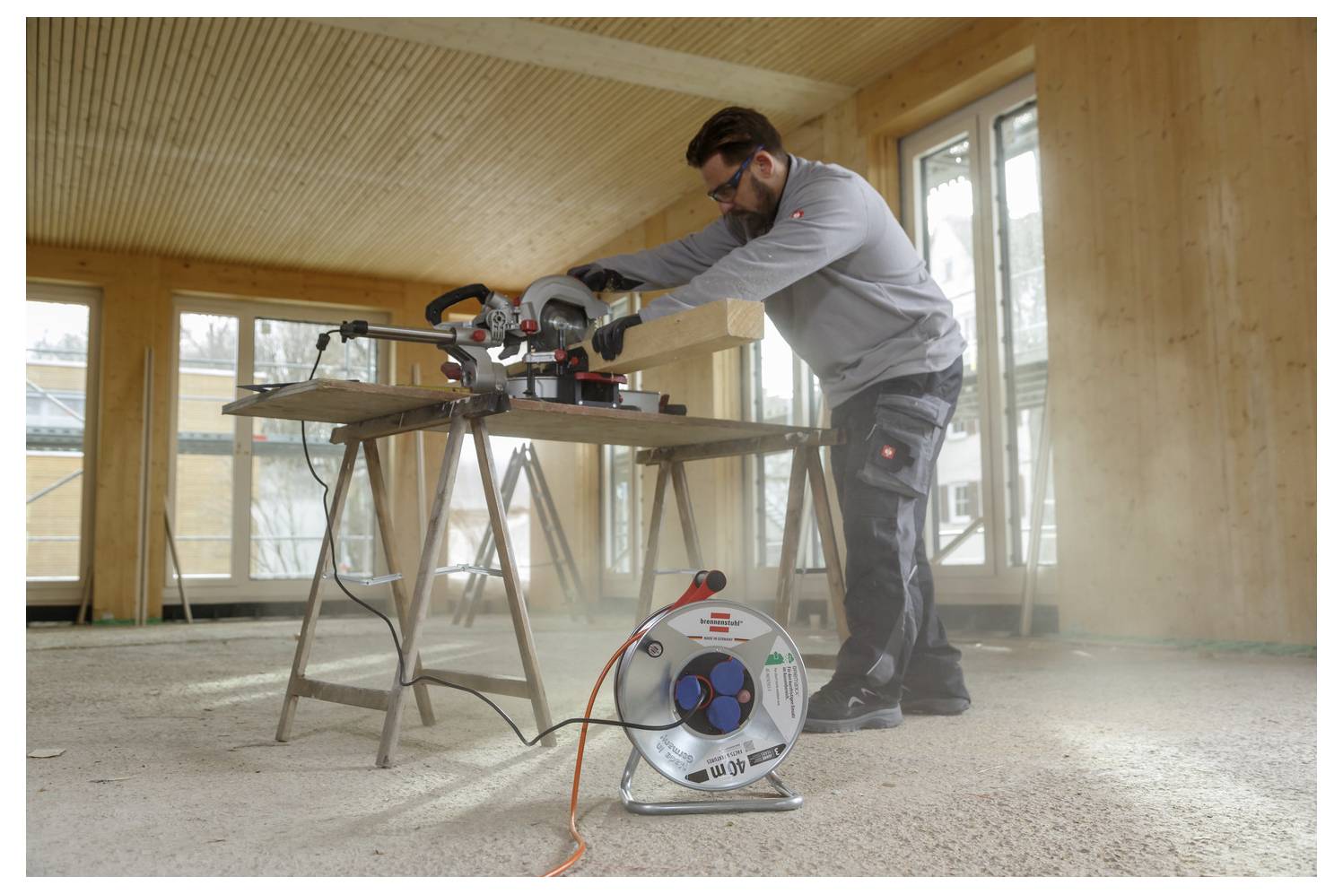 Tesar, ki dela s krožno žago na leseni deski pri delovni mizi v nedokončani sobi; kolut z navojno vrvjo je na tleh v bližini.