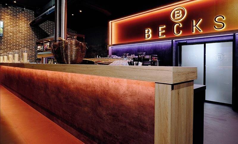 Slogovna bar z moderno osvetlitvijo. Neonski svetlobni napis z napisom 'BECKS'. Leseni šank v ospredju; stekleni izdelki v ozadju.