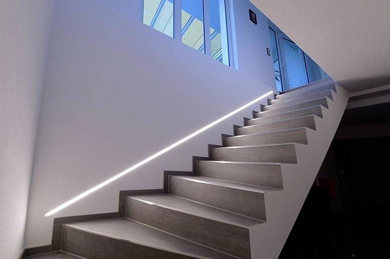 Svetla stopnišče z moderno osvetlitvijo v ograji, ki vodi do modre steklene vrat. Strešna okna zgoraj poskrbijo za naravno dnevno svetlobo.