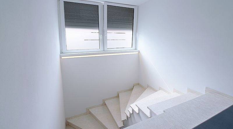 Bela stopnišče vodi do okna z zaprtimi roletami, ki skozi prostor razprostira razpršeno svetlobo. Minimalistični dizajn.