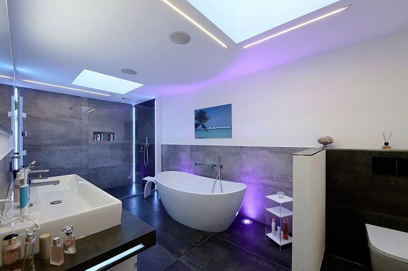 Moderno kopalnico s prosto stoječo kadjo, dvema umivalnikoma in LED-osvetlitvijo. Temne ploščice in strešno okno ustvarjajo elegantno atmosfero.