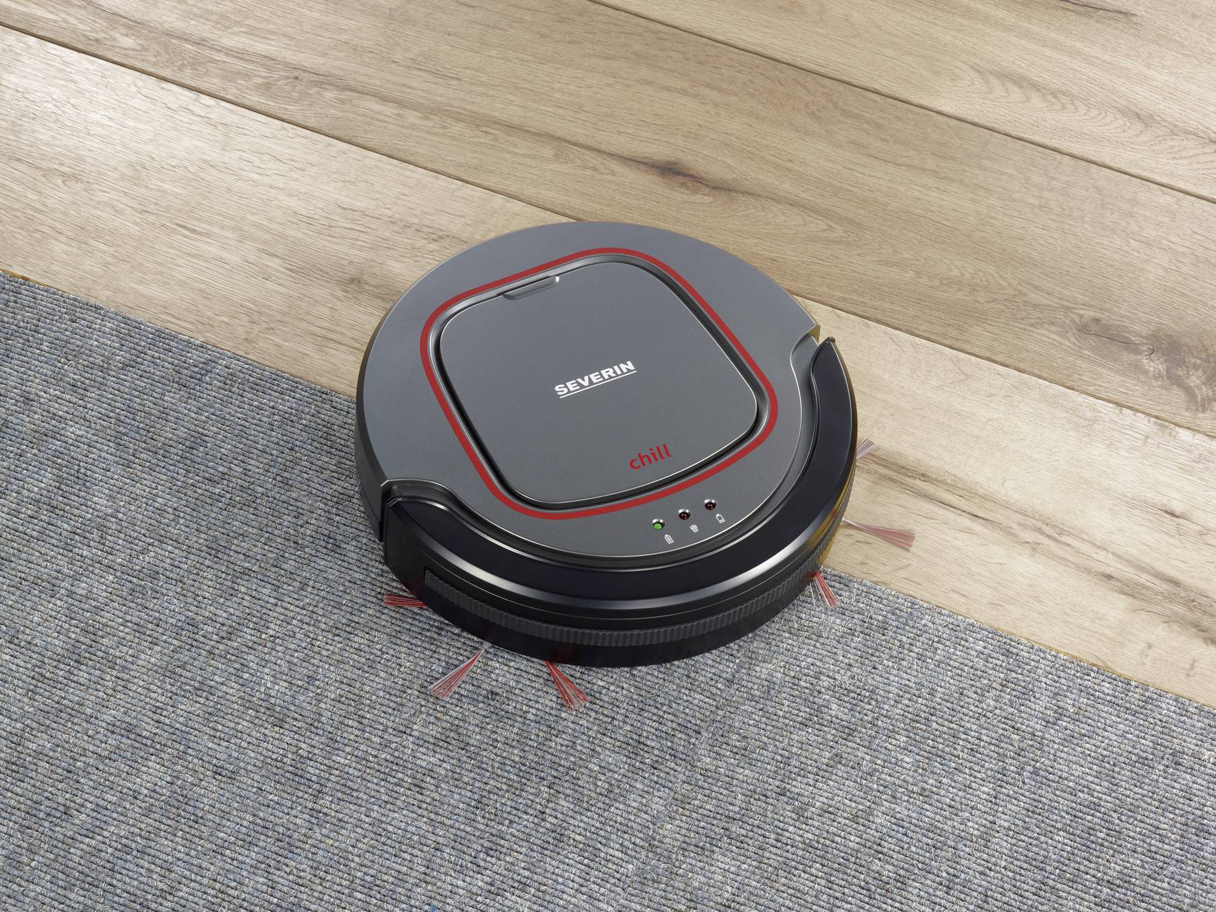 Okrogel črn sesalni robot čisti tla, prehaja med preprogo in lesenim podom. Enostavno upravljanje z gumbi na vrhu.