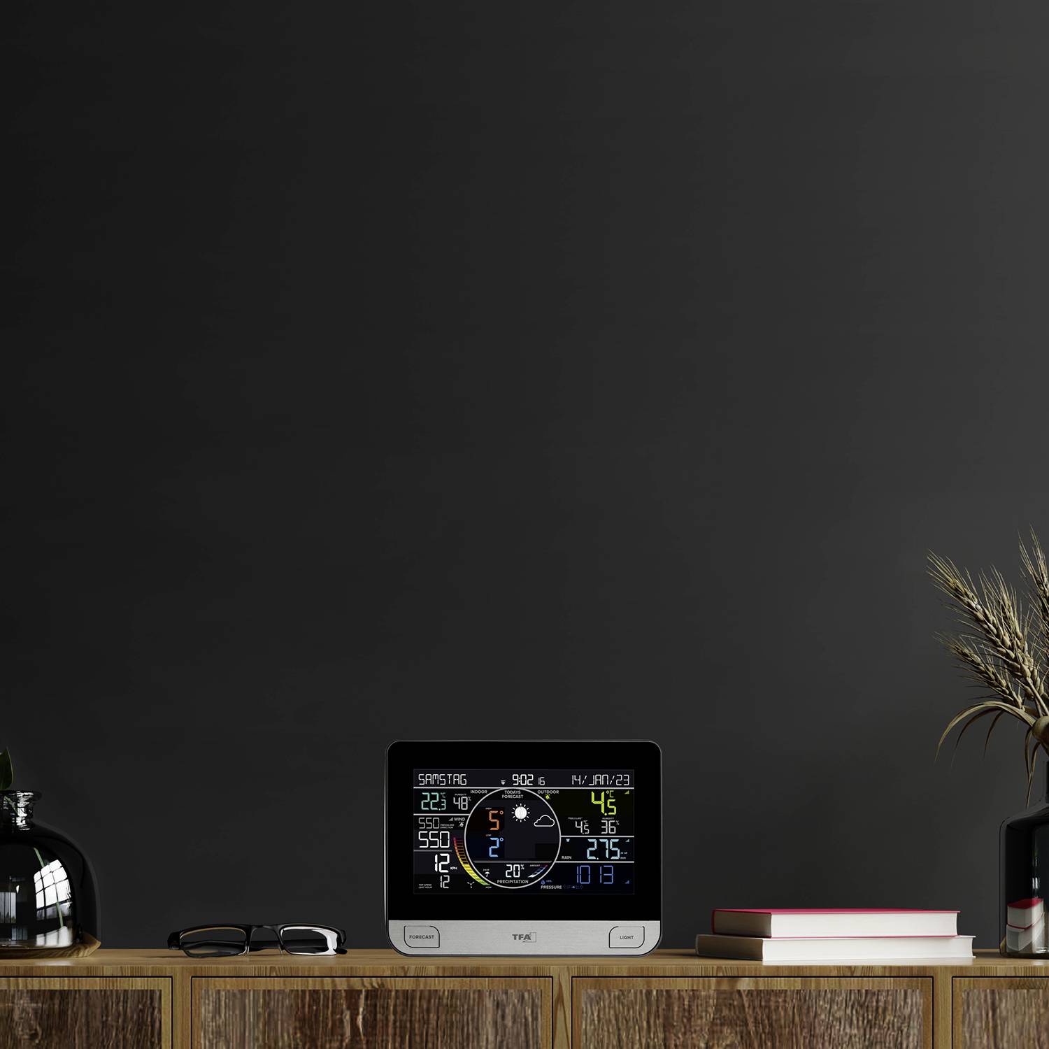 Vremenska postaja na polici prikazuje notranjo in zunanjo temperaturo, vlažnost zraka in zračni tlak. Poleg nje sta dekorativna vaza in knjige.