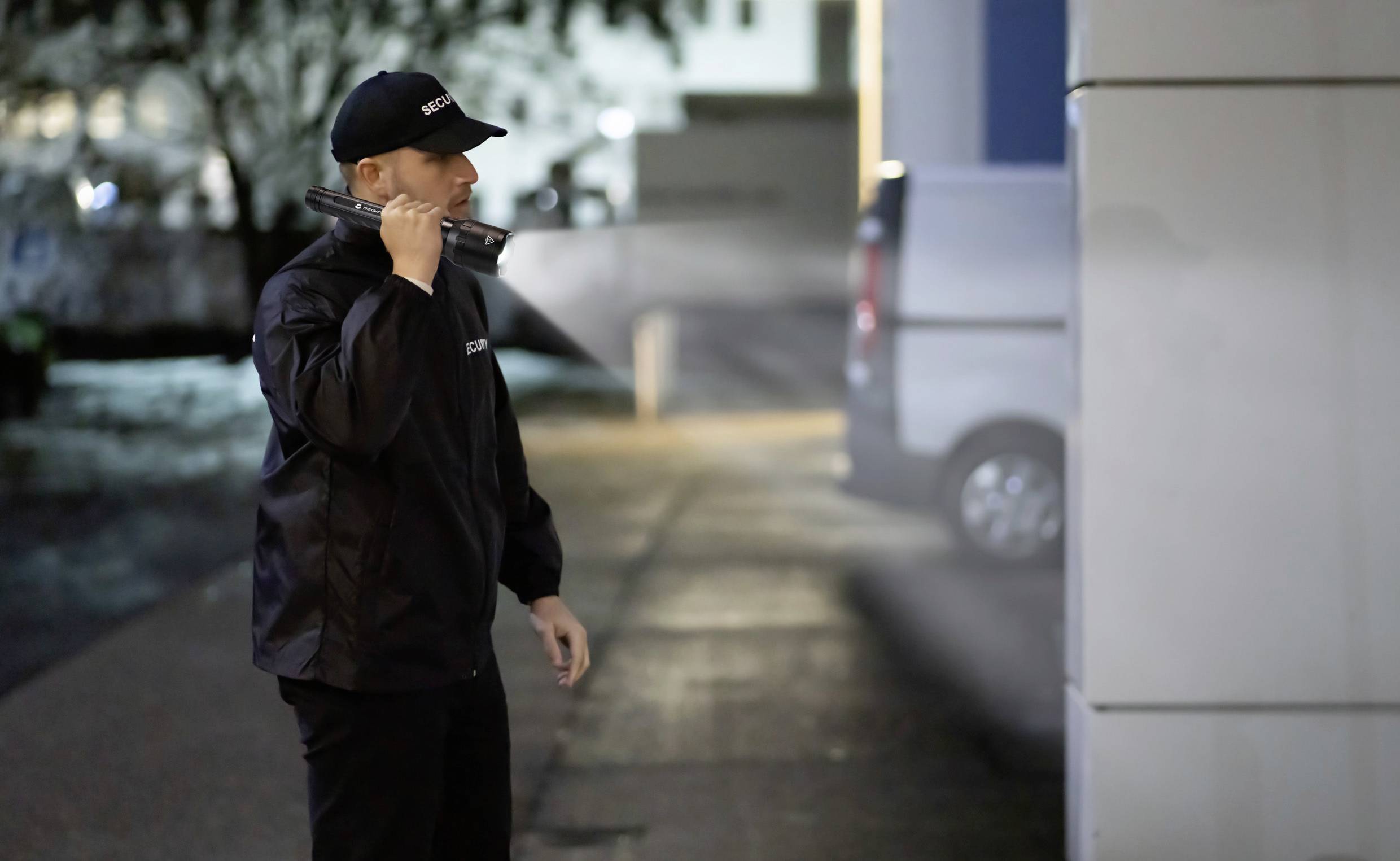 Varnostni agent v uniformi osvetljuje območje pred stavbo z žepno svetilko ponoči, medtem ko pozorno opazuje.