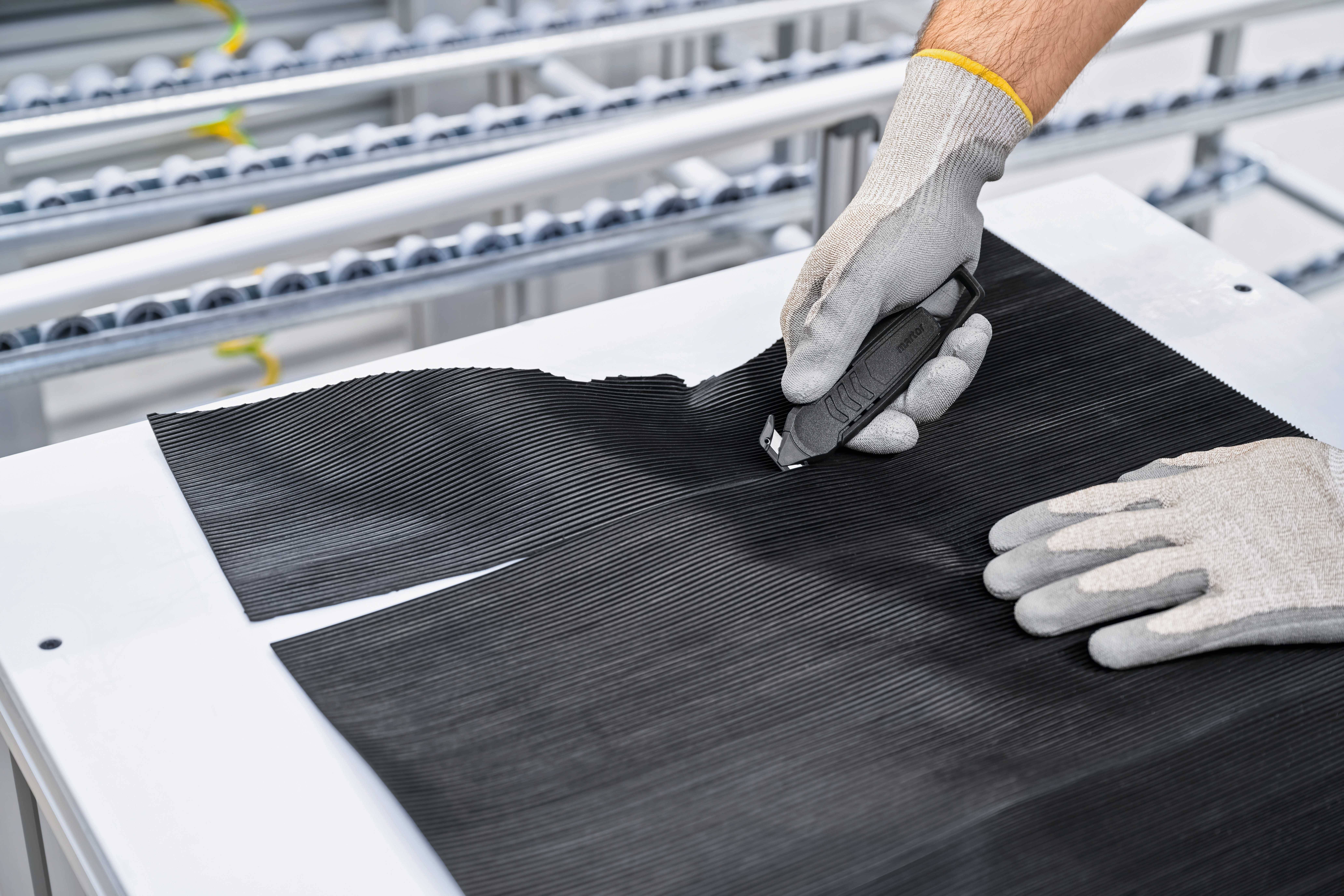 Oseba z nožem reže črn košček materiala na beli mizi. Nosi rokavice. V ozadju so vidni strojni elementi.
