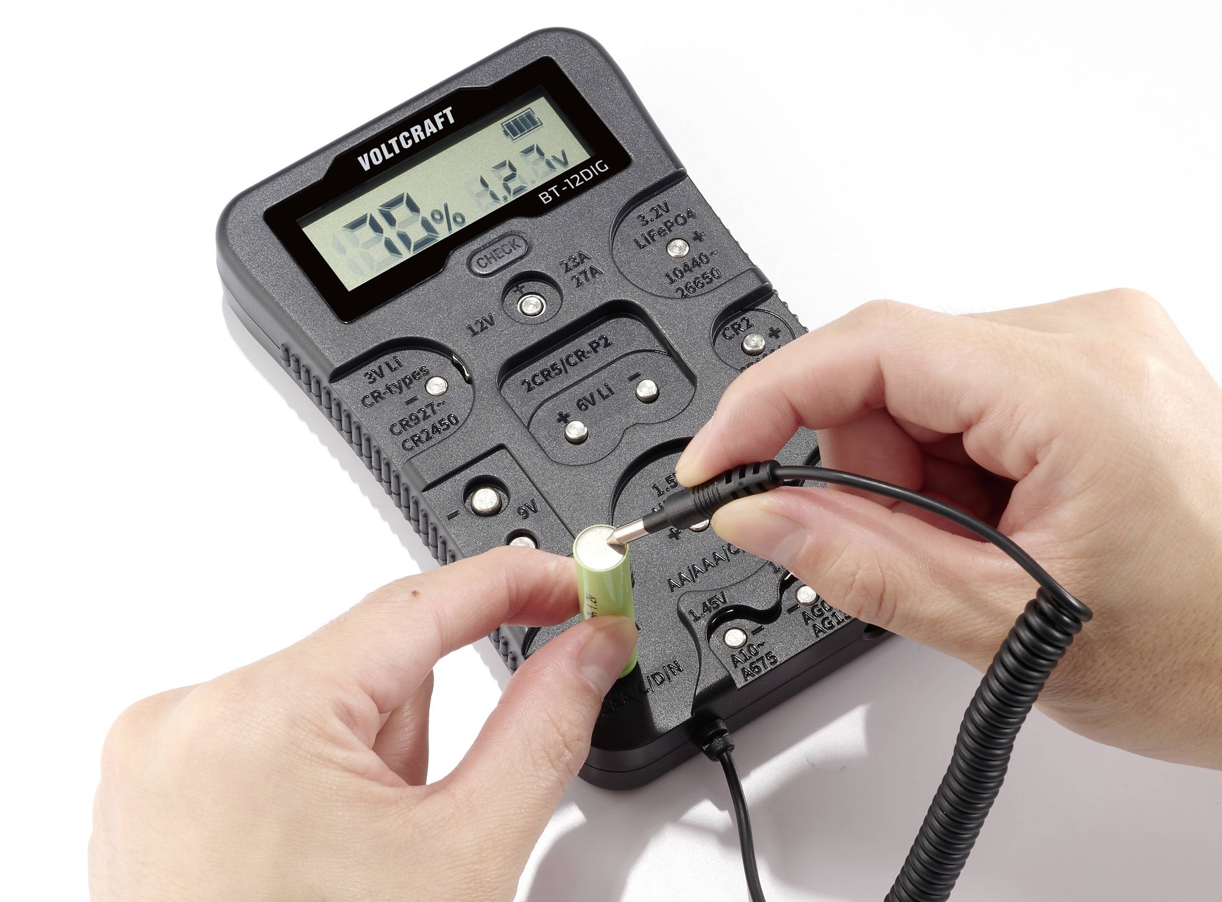 Oseba drži multimeter in meri valjasto baterijo. Naprava na zaslonu prikazuje izmerjeno vrednost.