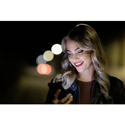 Nasmešena ženska z belimi lasmi gleda v svoj pametni telefon, medtem ko stoji zunaj ponoči. V ozadju svetijo luči.