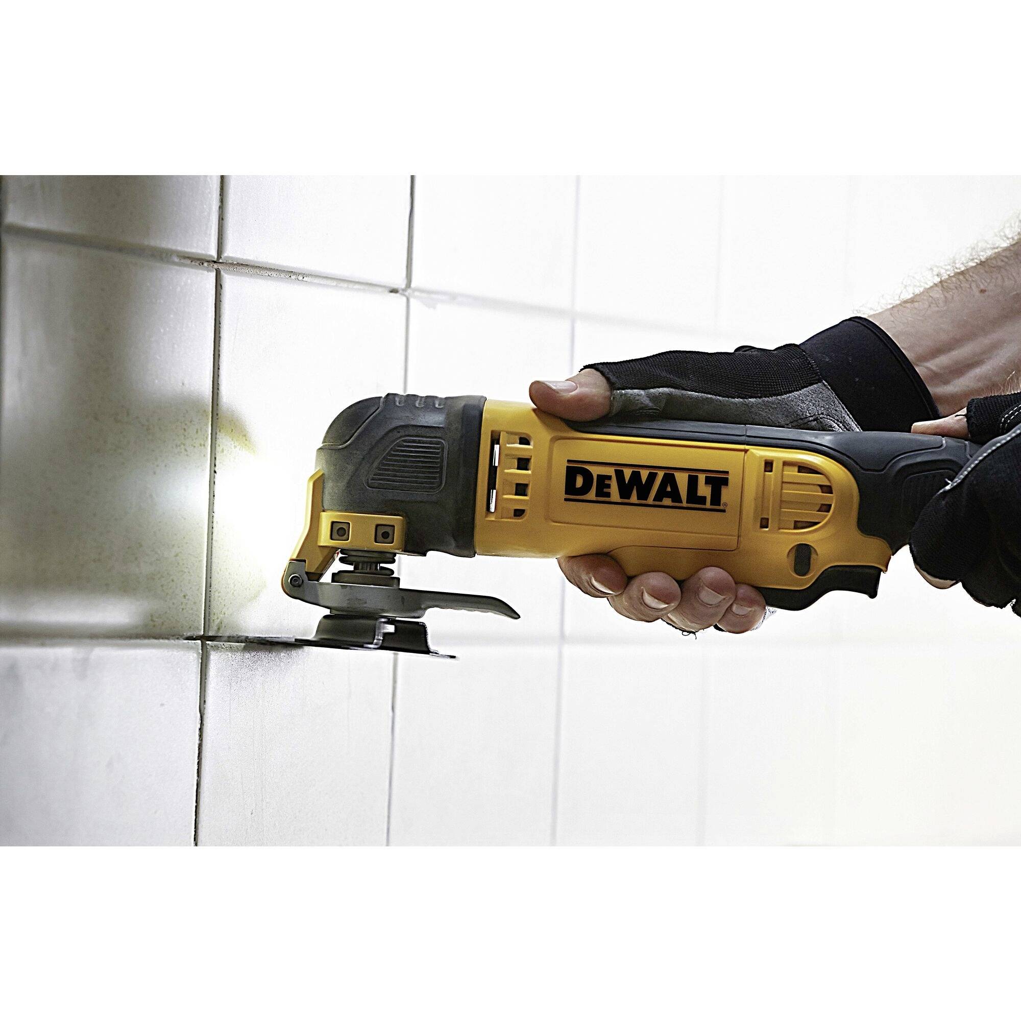 Ena roka drži električno večnamensko orodje znamke DeWalt in reže fuge na belo ploščičasti steni.